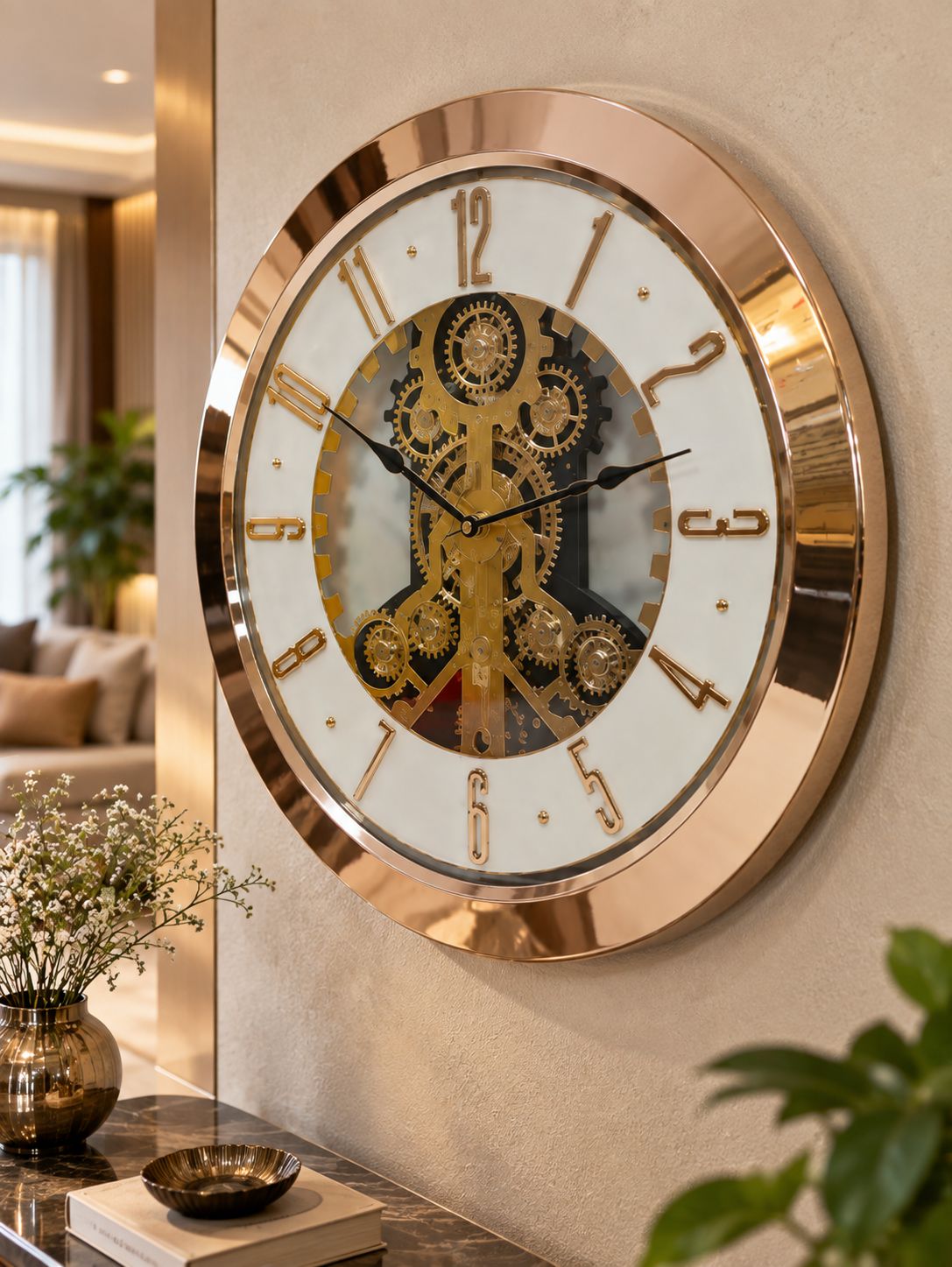 A large round wall clock with a rose gold metallic frame and a white face featuring a central gold mechanical gear design, mounted on a beige wall in a modern living room setting.