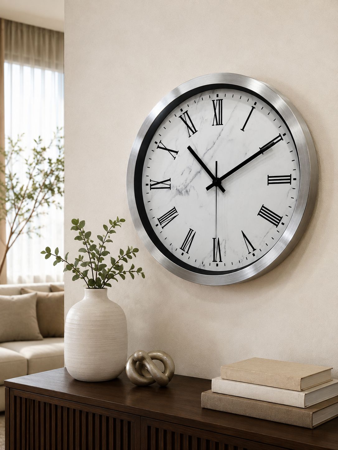 A large round wall clock with a silver metal frame and a white marble-pattern dial featuring black Roman numerals, mounted on a beige wall in a modern living room setting.