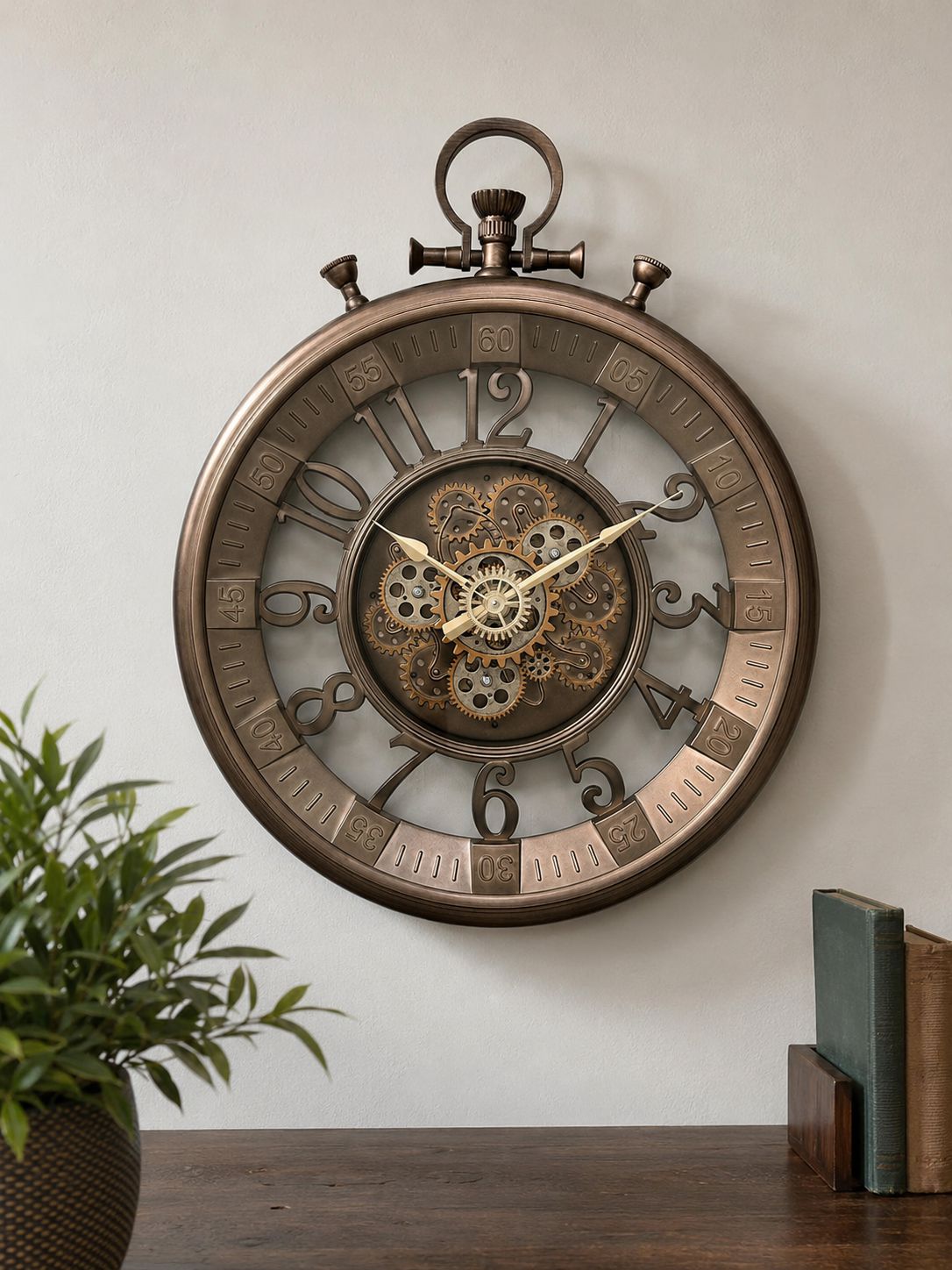 A large round wall clock with a vintage stopwatch design, featuring a bronze-toned metal frame, exposed internal gears, and large Arabic numerals, mounted on a light wall above a wooden surface with a plant and books.