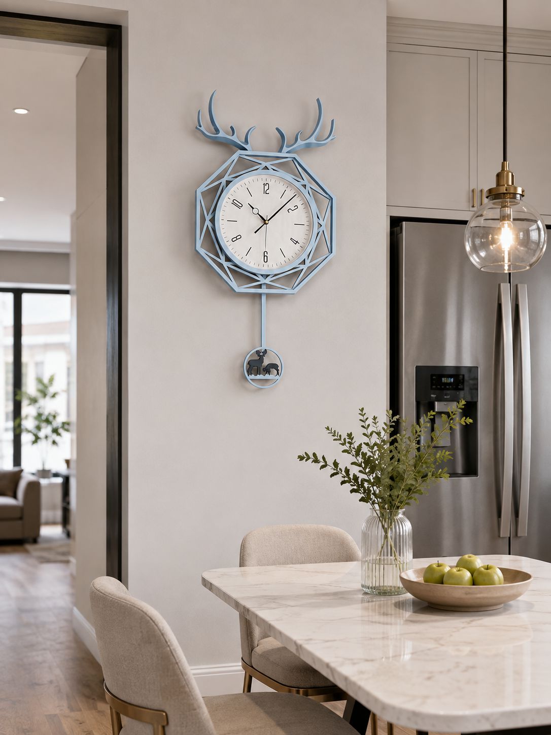 A light blue geometric wall clock with antler details hangs on a beige wall in a modern kitchen setting, with a marble dining table, chairs, and a stainless steel refrigerator visible.