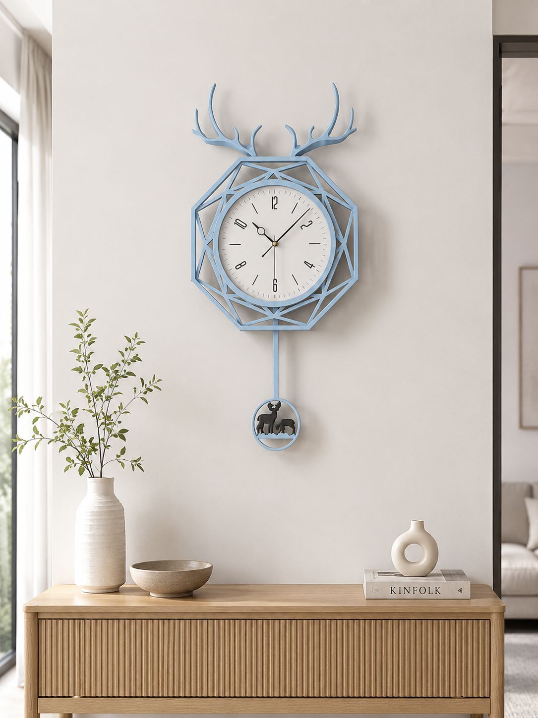 A light blue geometric wall clock with antler motifs and a hanging bear pull, displayed on a beige wall above a wooden console table with a vase and books.