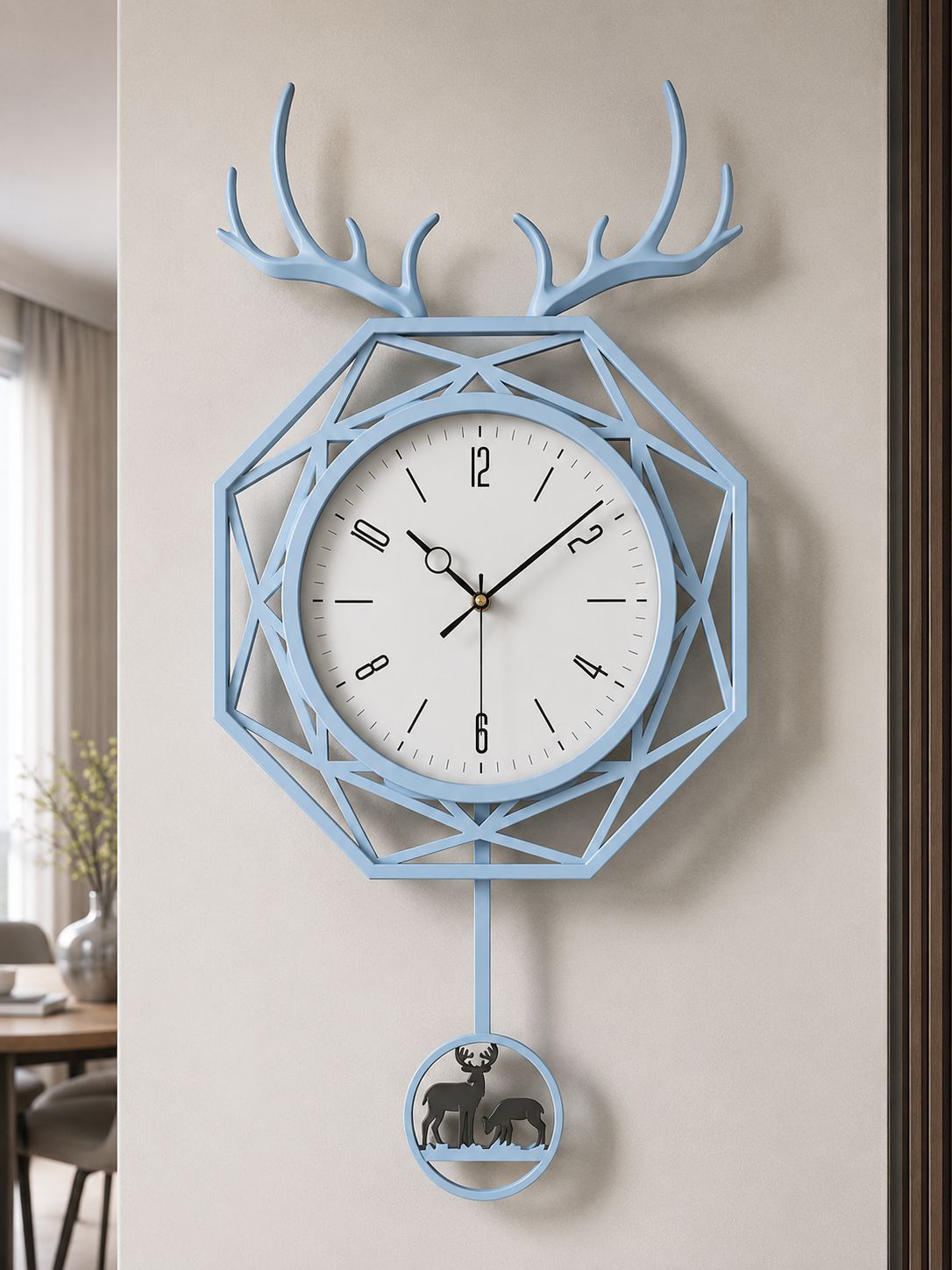 A light blue geometric wall clock with antler motifs at the top and a hanging circular ornament below, shown mounted on a beige wall in an indoor room setting.