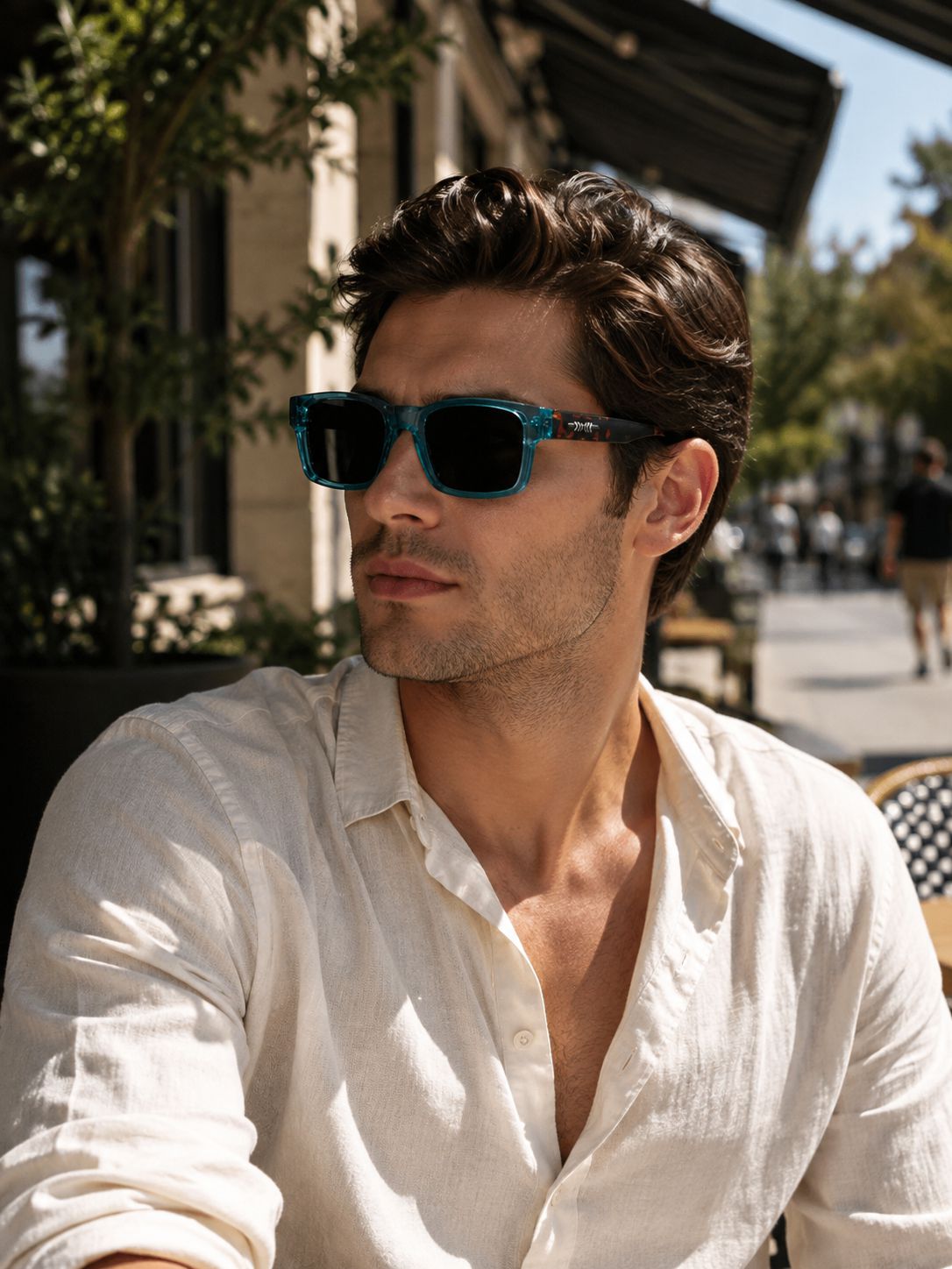 A man wearing blue-framed sunglasses with dark lenses and a white linen shirt, photographed outdoors on a sunny street or patio with blurred buildings and trees in the background.