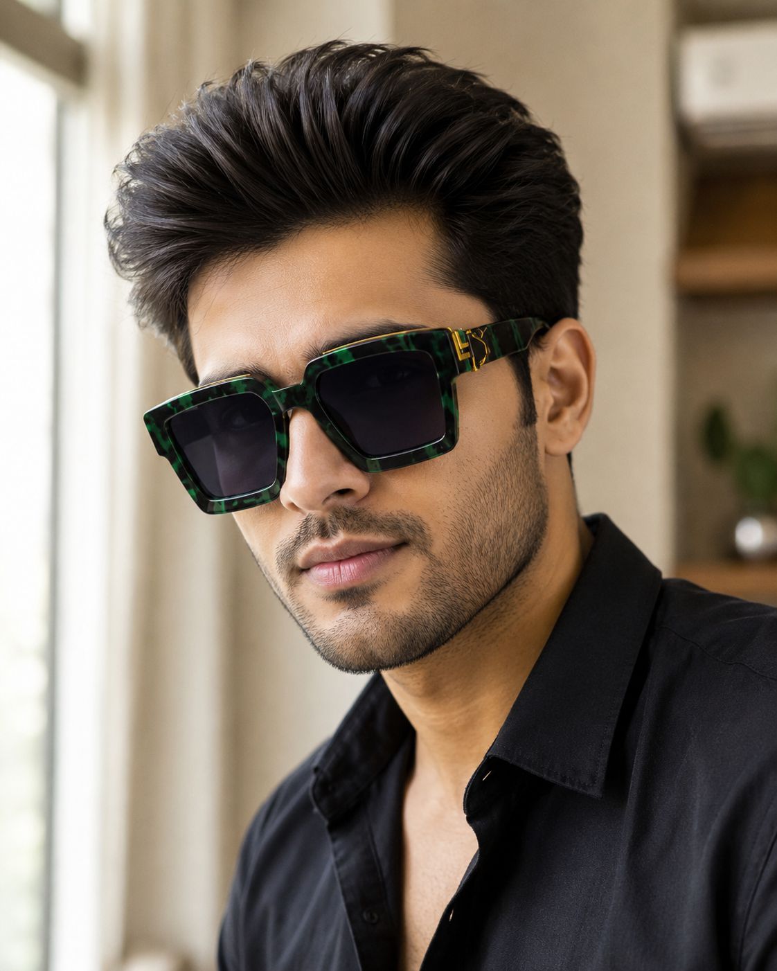 A man wearing green tortoiseshell square-frame sunglasses with dark lenses and a gold accent on the temple, paired with a black shirt in a softly lit indoor setting.