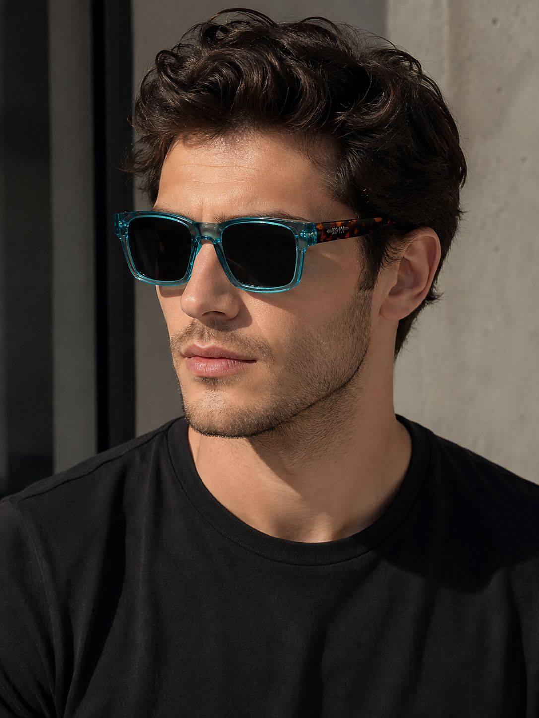 A man wearing square sunglasses with a translucent teal-blue frame and dark lenses, paired with a black t-shirt against a concrete wall.