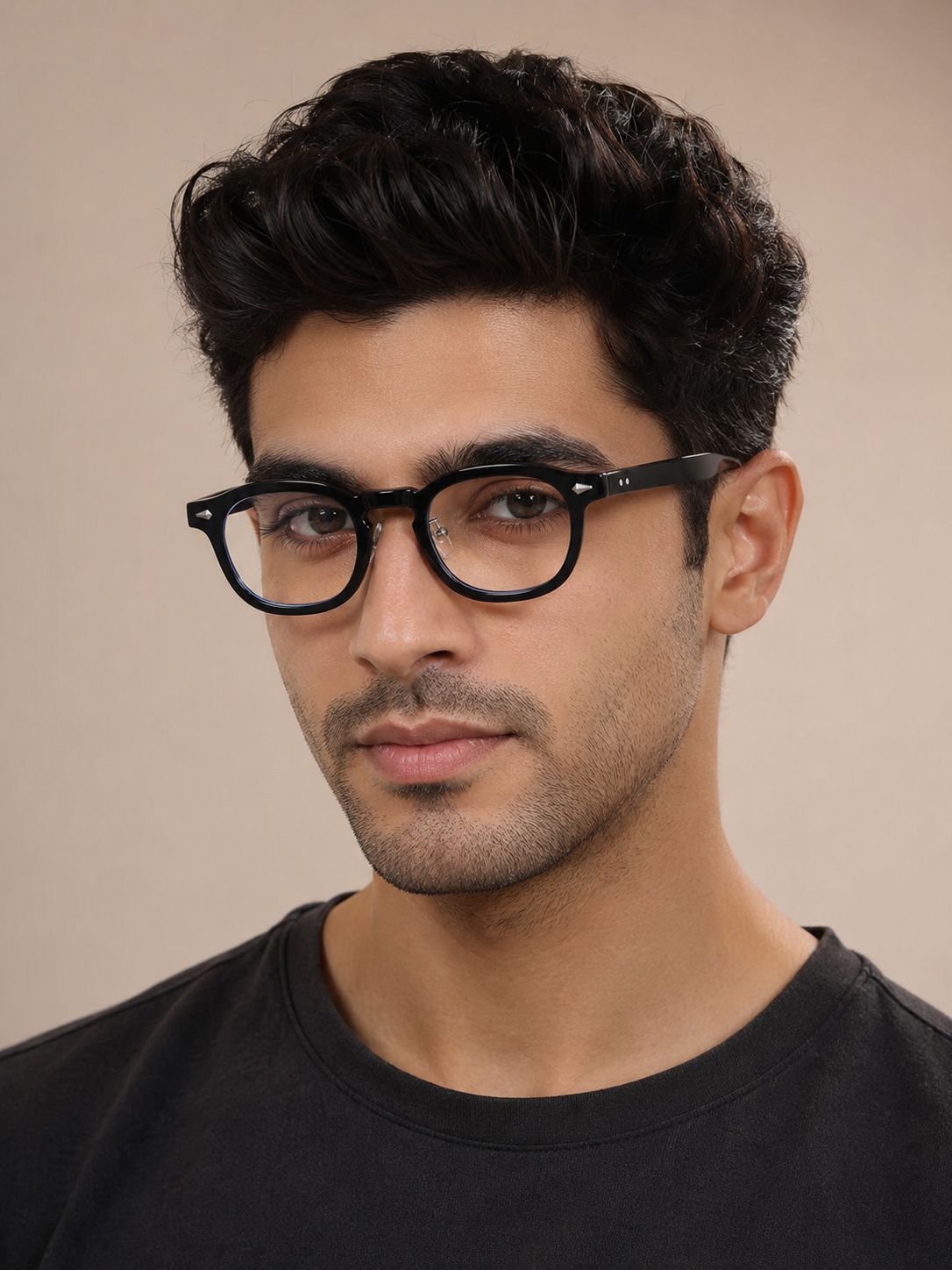 A man with styled dark hair wearing black round-frame eyeglasses and a black crew-neck t-shirt, photographed against a solid beige background.