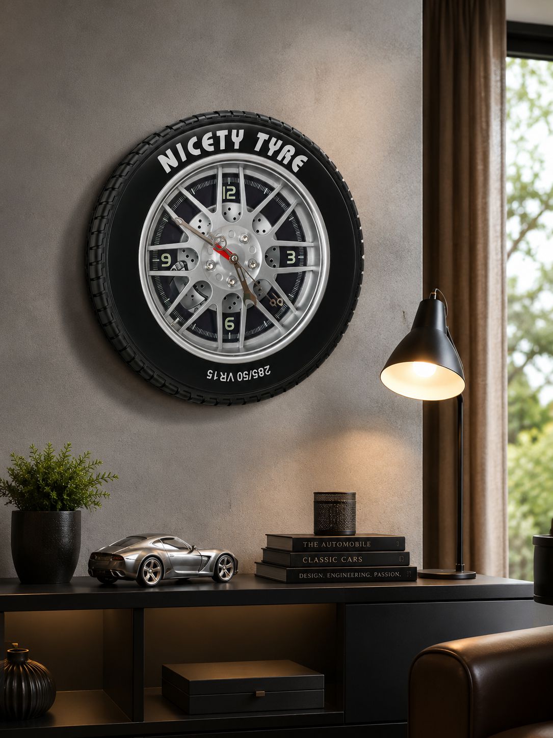 A novelty wall clock designed to look like a black car tire with a silver multi-spoke wheel and clock face in the center, mounted on a textured gray wall in a modern room setting.