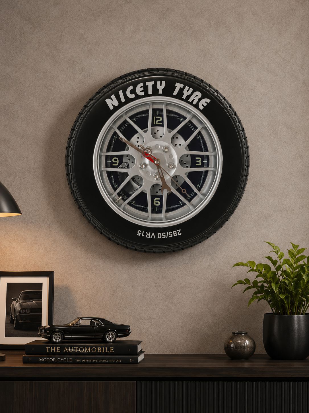 A novelty wall clock shaped like a black tire with a silver and white multi-spoke wheel center and white 'NICETY TYRE' text, mounted on a textured beige wall above a dark console table with decor.