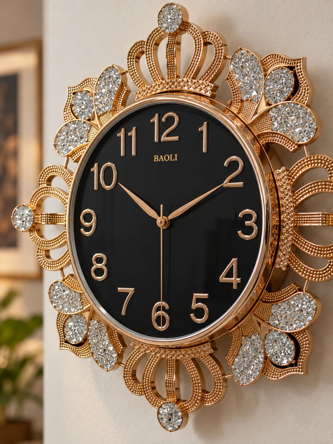 A ornate wall clock with a black dial and a highly decorative gold-tone frame embellished with clear crystals, mounted on a light beige wall.