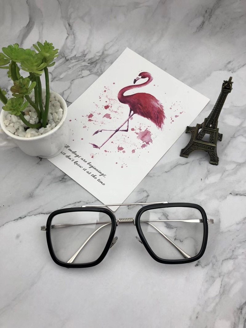 A pair of Tony Stark Avengers End Game sunglasses with square frames, displayed on a marble surface next to a paper with a flamingo drawing and a small Eiffel Tower figurine.