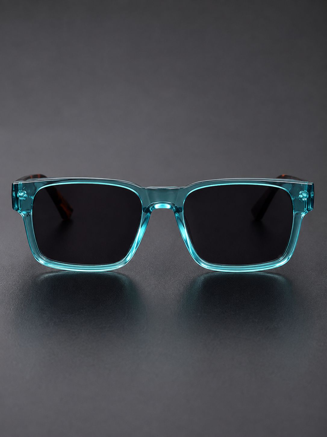 A pair of square sunglasses with translucent aqua blue frames and dark lenses, displayed on a dark gray reflective surface.