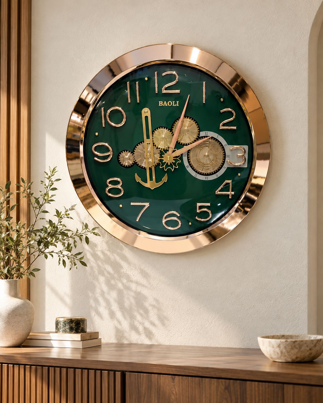 A round decorative wall clock with a dark green face, gold-tone numerals and hands, exposed gear details, and a metallic gold frame, mounted on a beige wall above a wooden console table with a plant and bowls.