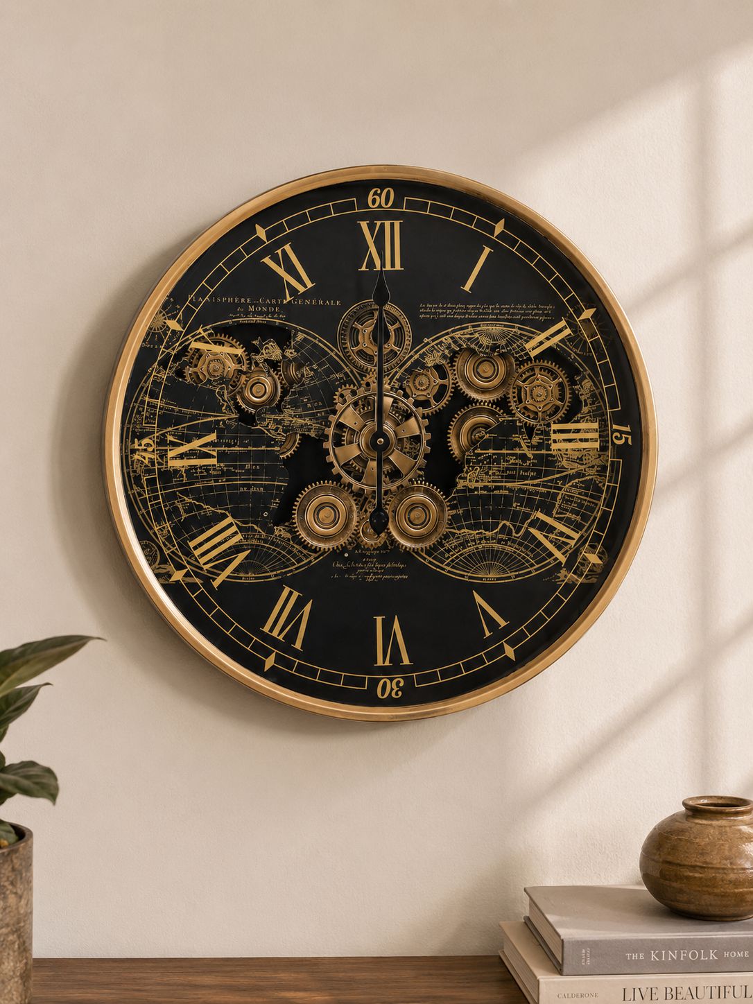 A round steampunk-style wall clock with a black face, gold Roman numerals, intricate gear illustrations, and a gold-toned rim, mounted on a beige wall above a wooden surface with books and a small vase.