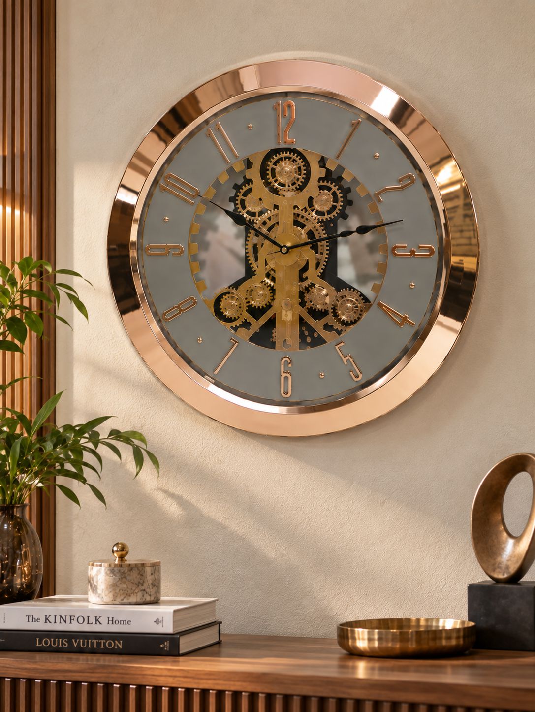 A round steampunk-style wall clock with a rose gold metallic frame and a gray face featuring gold gears, mounted on a beige wall above a wooden console table with decorative objects.