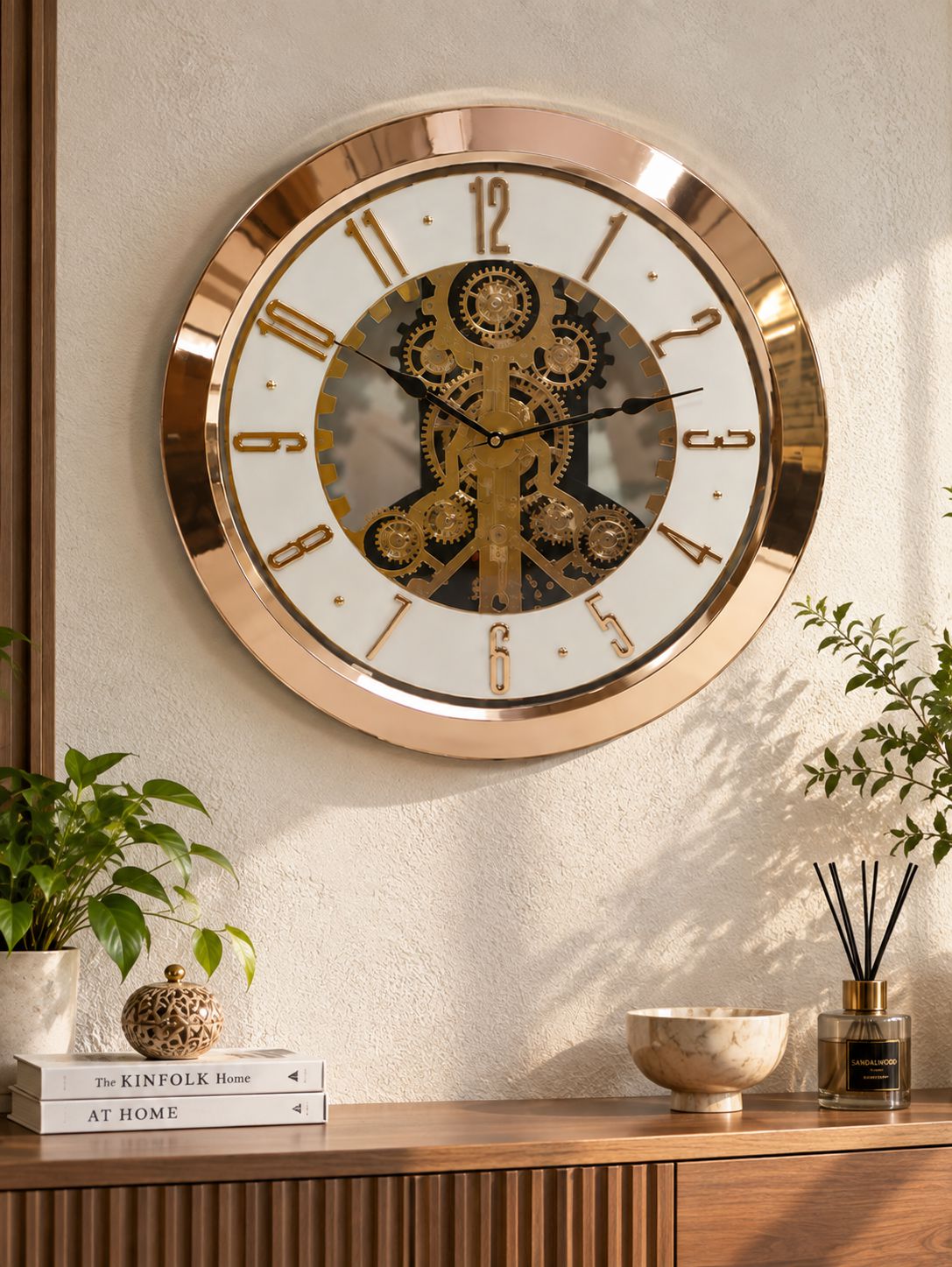 A round steampunk-style wall clock with a rose gold metallic frame, white face, black numerals, and a central gold gear mechanism, mounted on a textured beige wall above a wooden console table with plants and decor.