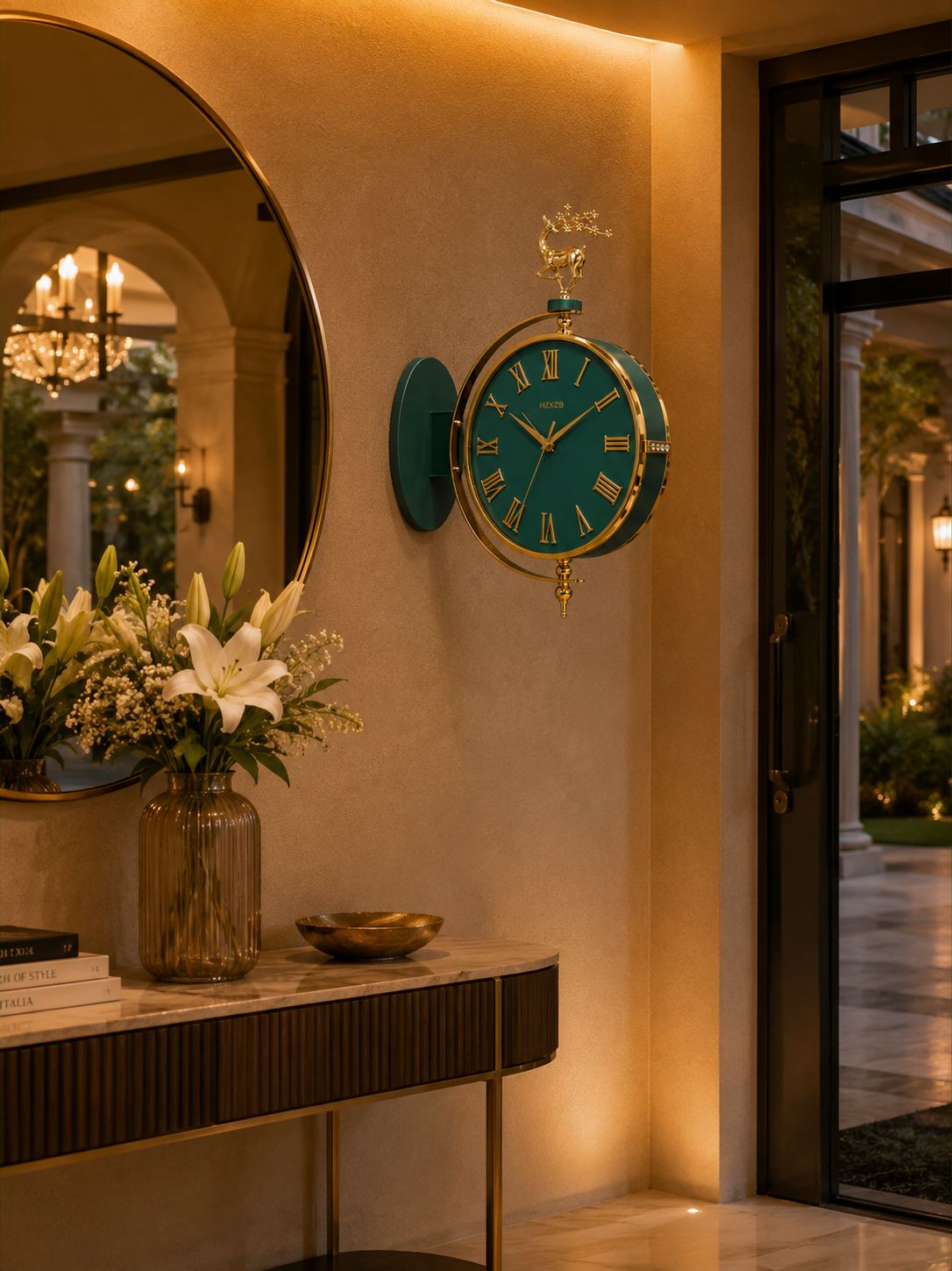 A round teal and gold wall clock with a deer topper, mounted on a warm beige wall in a luxury interior setting next to a large oval mirror and a console table with flowers.
