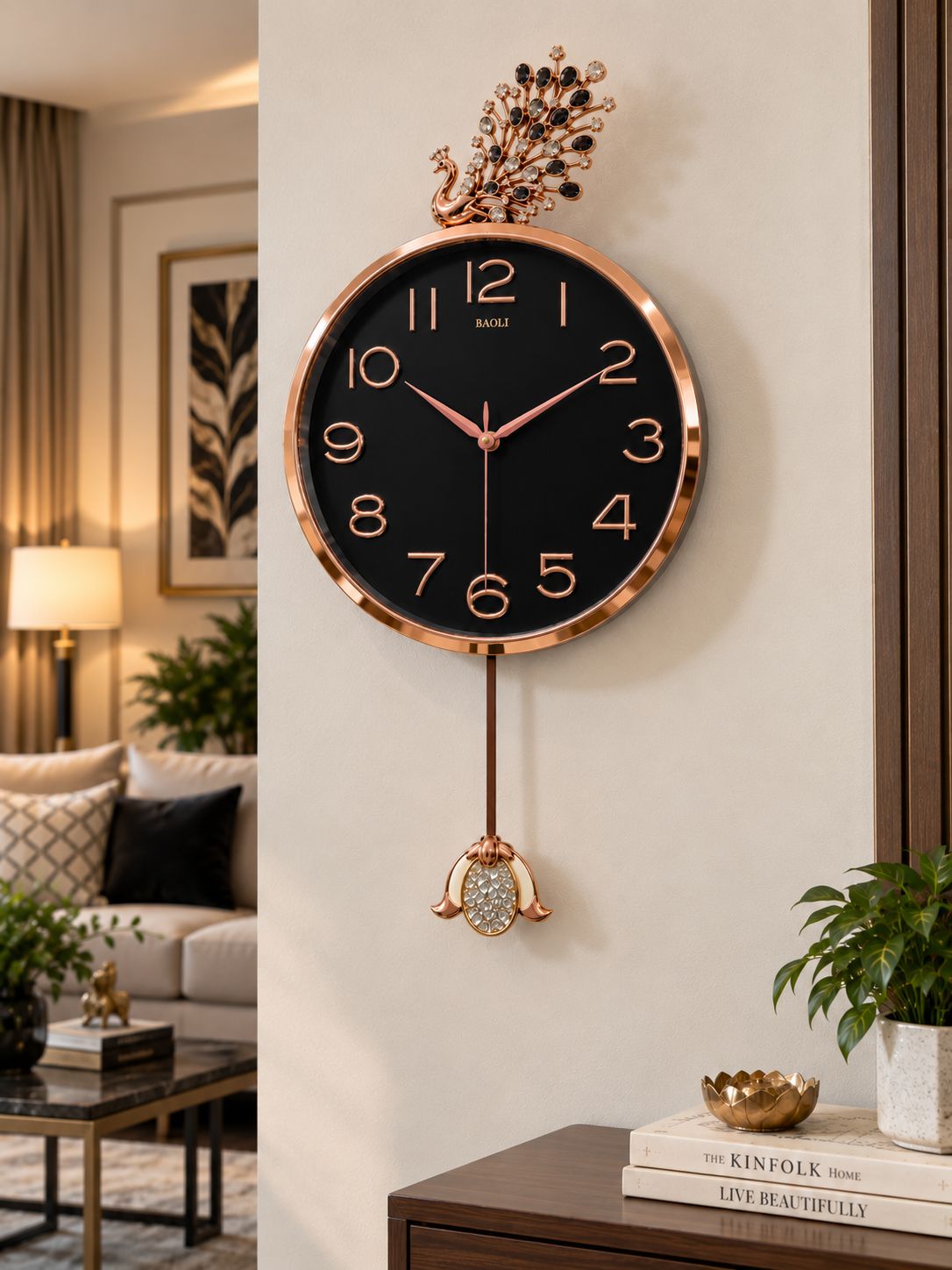 A round wall clock with a black face, rose gold-tone frame and numerals, and a decorative hanging ornament below, shown mounted on a beige wall in a styled living room interior.