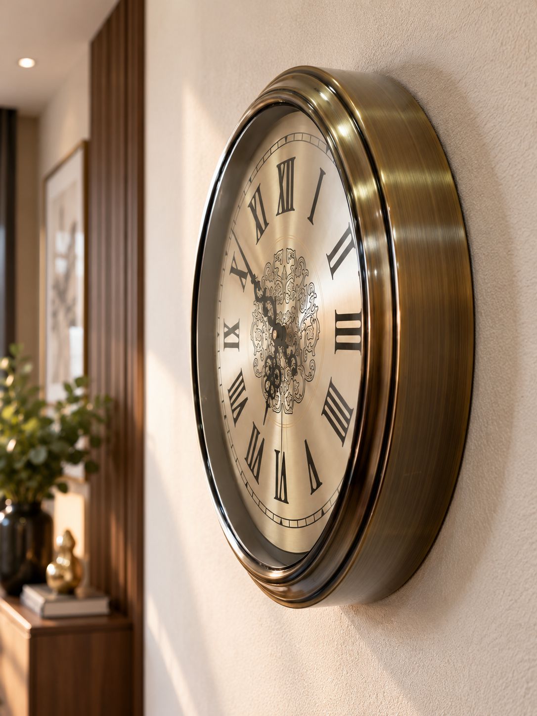 A round wall clock with a brushed brass frame and Roman numerals, mounted on a textured beige wall in a warm, modern interior setting.