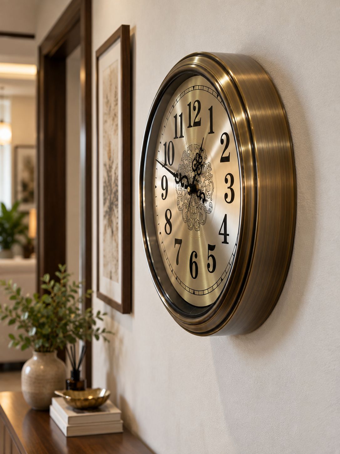 A round wall clock with a brushed brass/gold-toned metal frame and a cream-colored dial featuring black Arabic numerals, mounted on a light beige wall in a styled interior hallway.