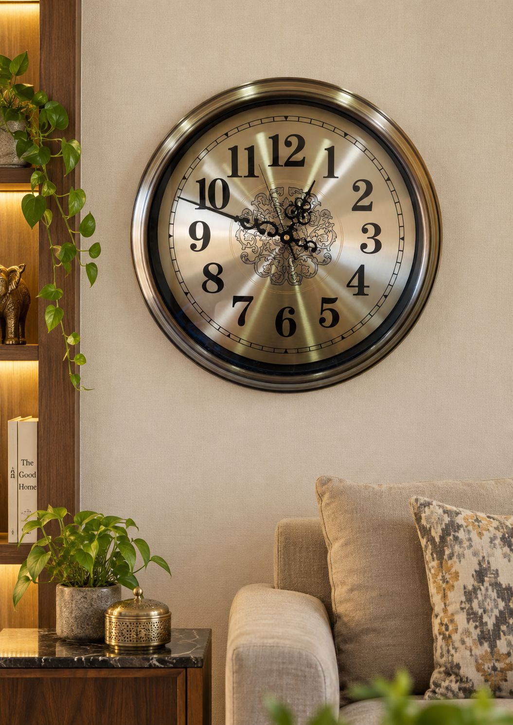 A round wall clock with a brushed gold-toned face and silver outer rim, mounted on a beige wall above a sofa in a styled living room setting.