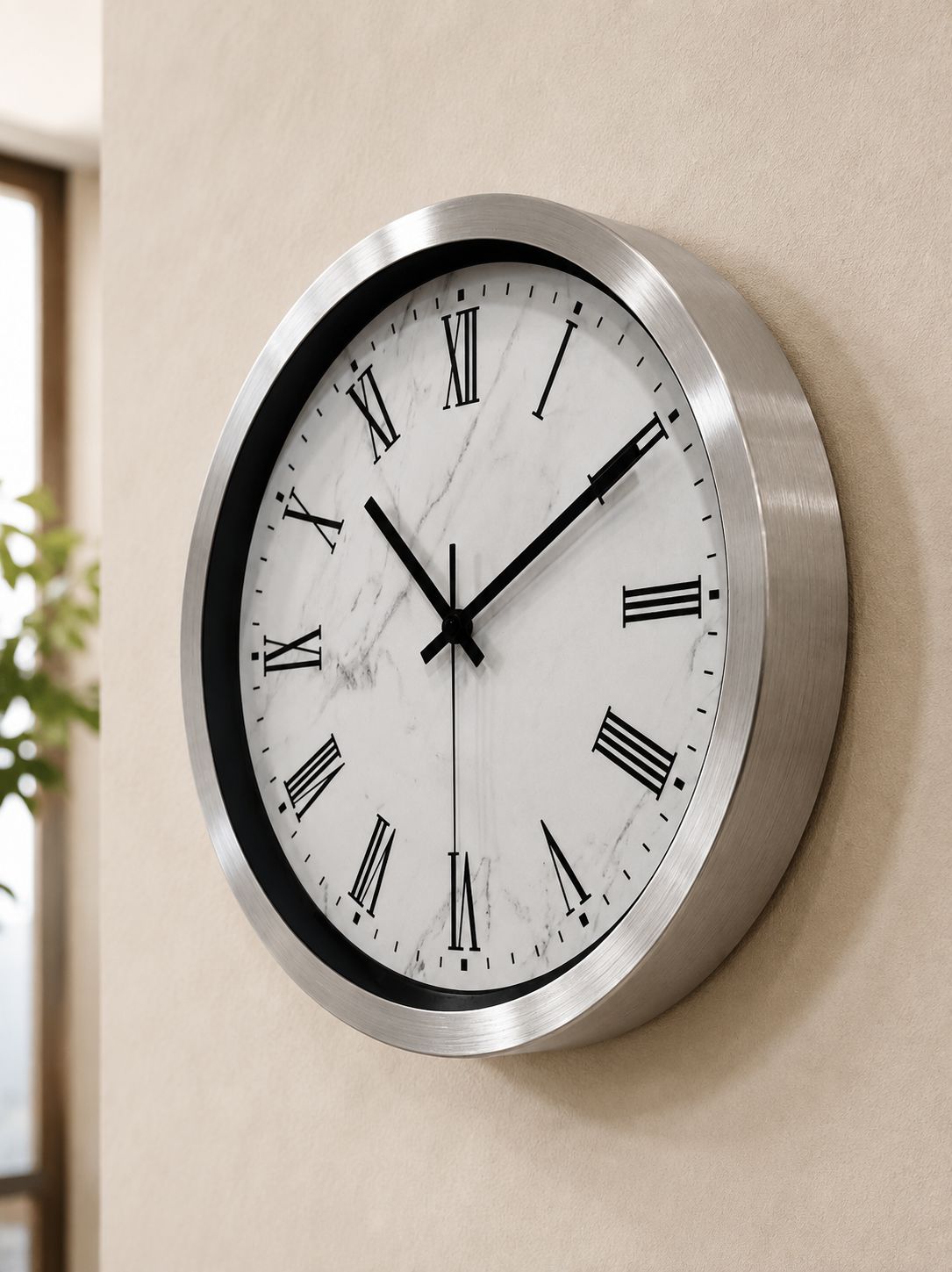 A round wall clock with a brushed silver metal frame, a white marble-patterned dial, and black Roman numerals, mounted on a beige wall.