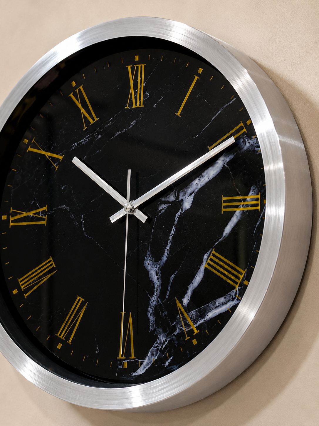 A round wall clock with a brushed silver metal frame and a black marble-pattern dial featuring gold Roman numerals and silver hands, mounted on a beige wall.