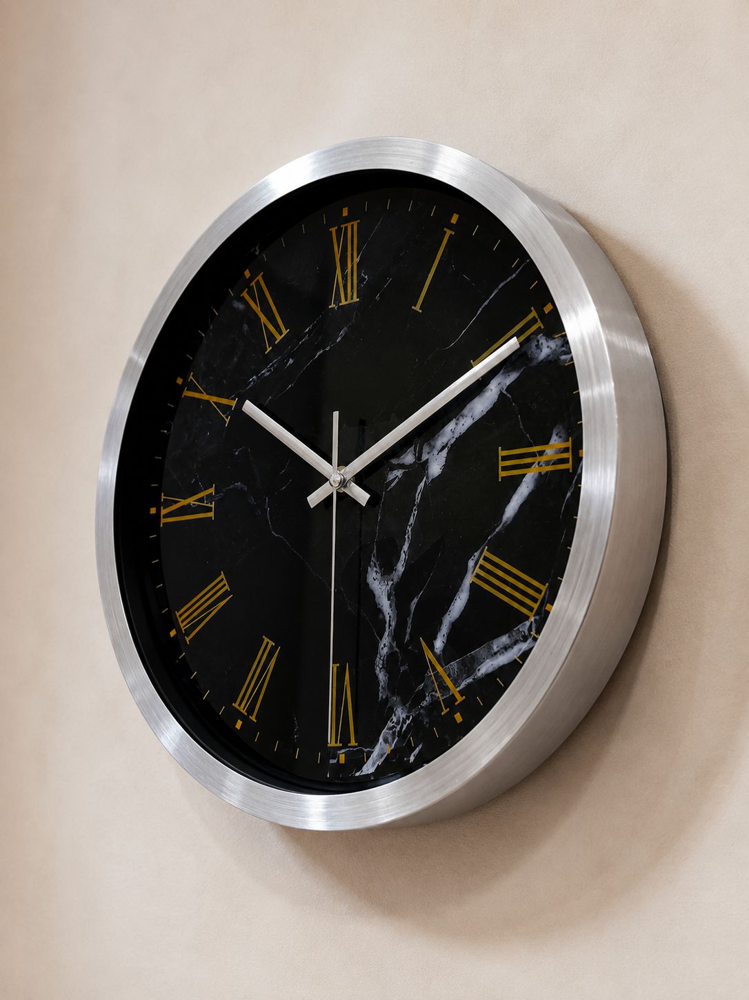 A round wall clock with a brushed silver metal frame and a black marble-pattern dial featuring gold Roman numerals, mounted on a beige wall.