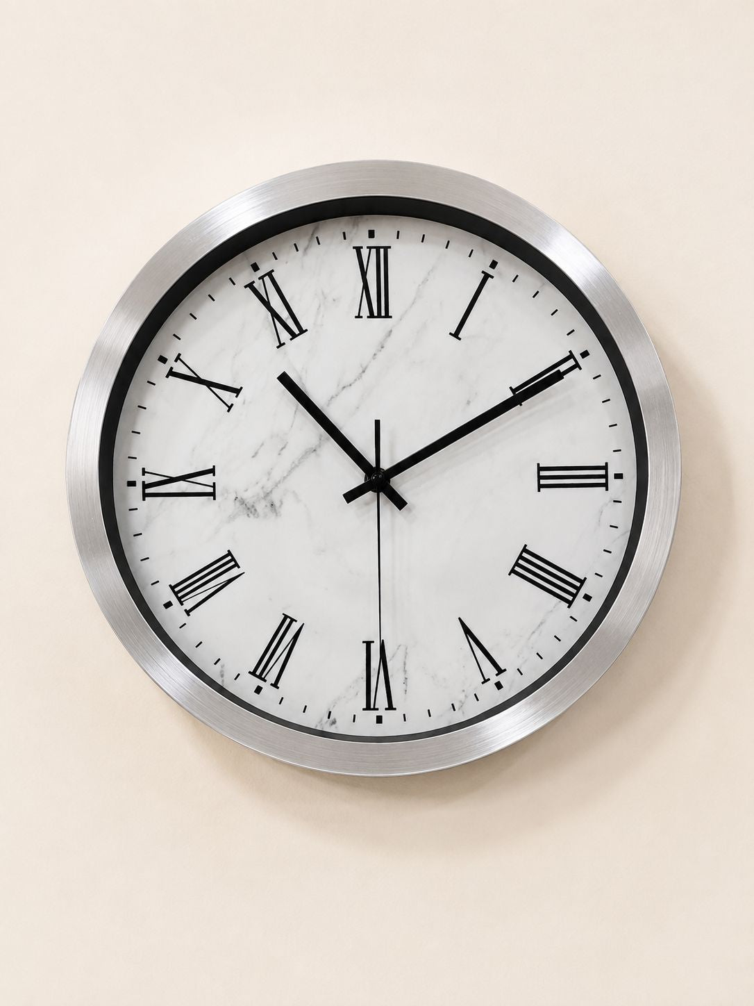 A round wall clock with a brushed silver metal frame, black hands, and black Roman numerals on a white marble-patterned face, mounted on a warm beige wall.