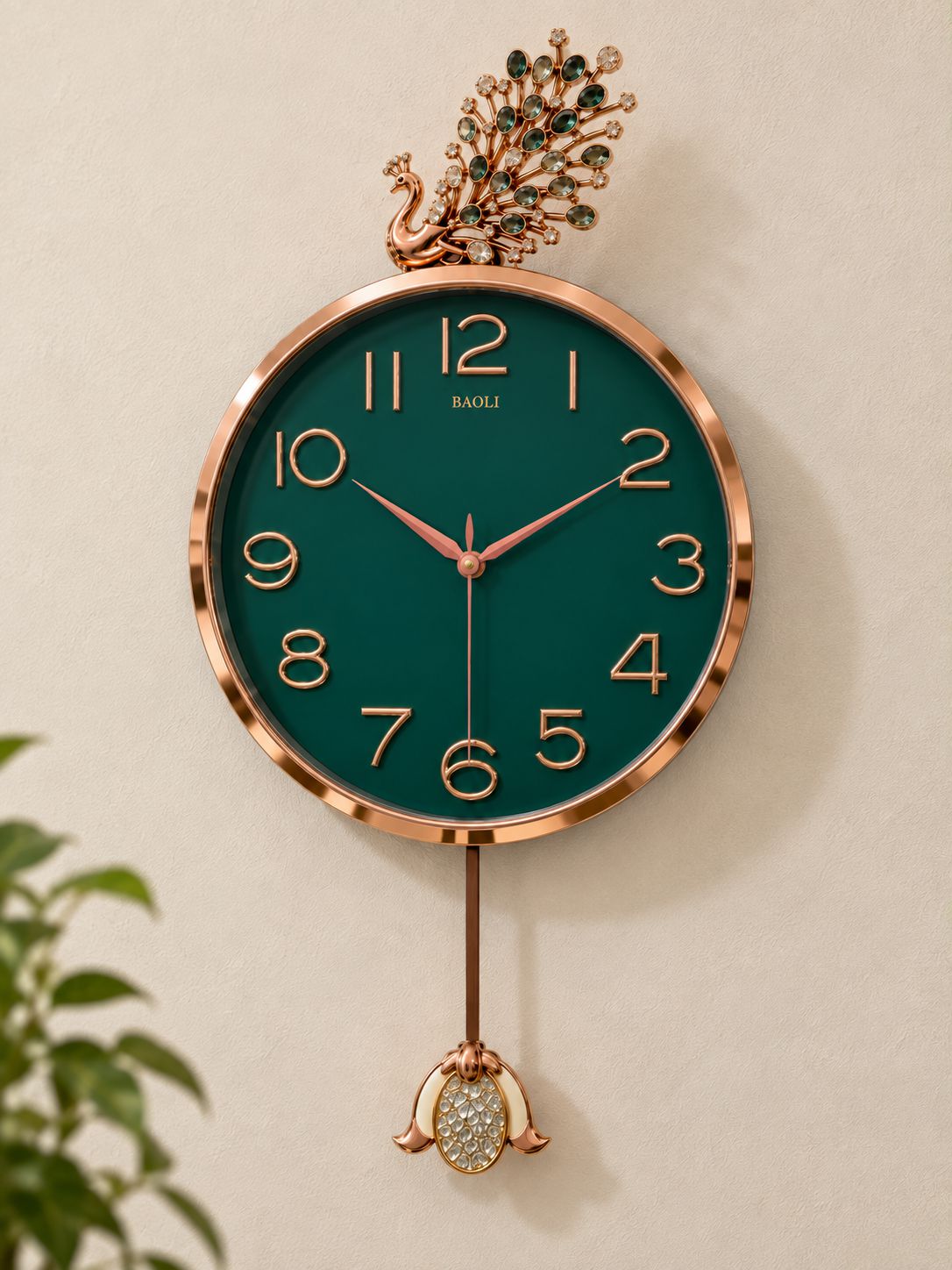 A round wall clock with a dark green dial and rose gold-toned frame, featuring a decorative peacock finial at the top and a dangling ornament at the bottom. The brand name 'BAOLI' is printed above the 12 o'clock mark.