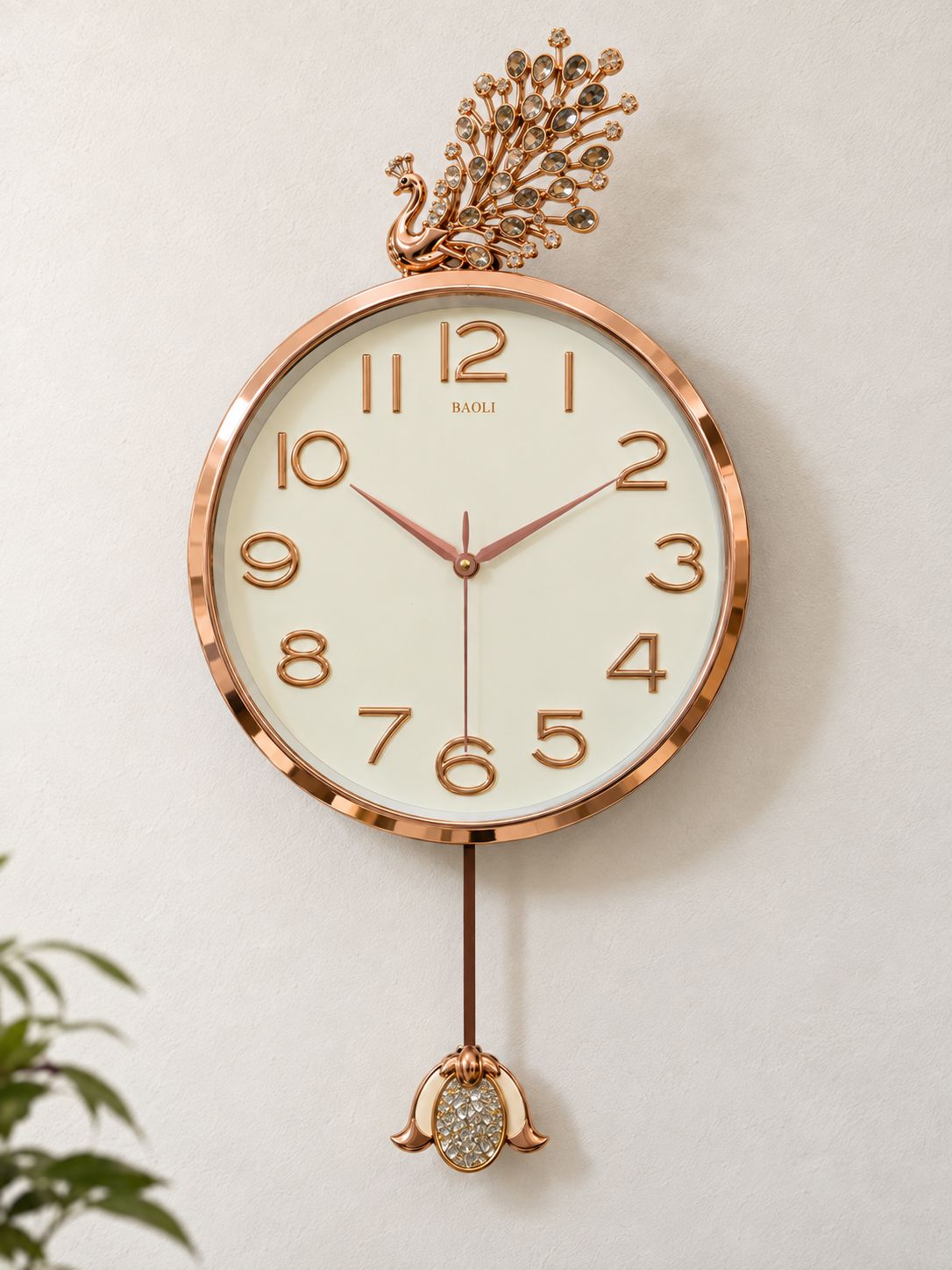 A round wall clock with a rose gold metallic frame and a cream-colored dial, featuring a decorative peacock topper and a dangling peacock weight with a rhinestone-encrusted center, mounted on a light beige wall.