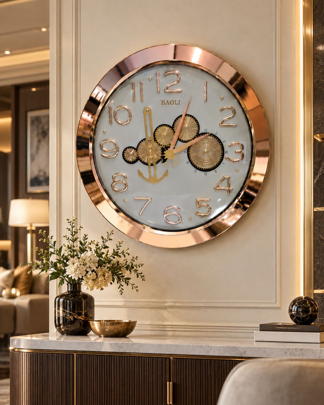 A round wall clock with a rose gold metallic frame and a light blue dial, featuring gold-tone numerals and decorative gear-like hands. The clock is mounted on a cream-colored wall in a modern, upscale interior setting with a marble console table and decorative vase below.