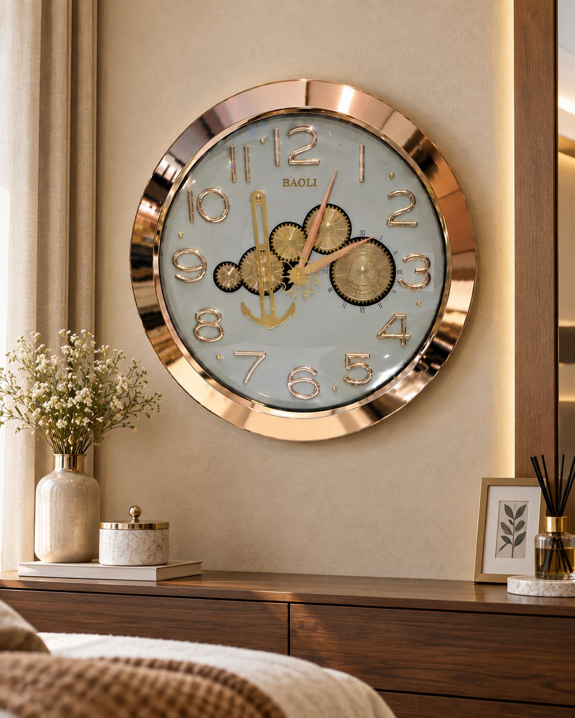 A round wall clock with a rose gold metallic frame and a light blue dial, featuring gold gears and an anchor motif. The clock is mounted on a beige wall above a wooden console table decorated with a vase of white flowers and other home accessories.