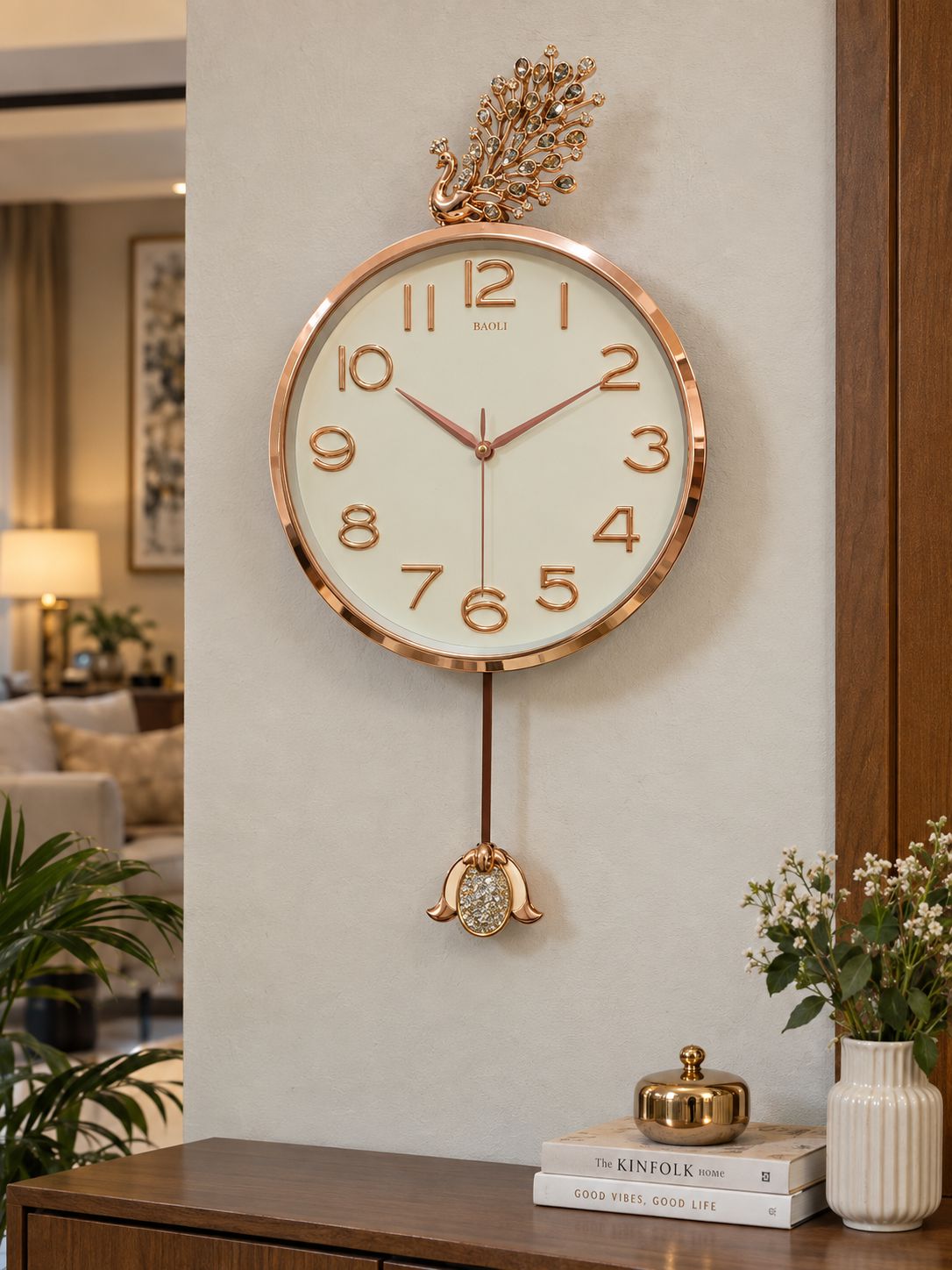 A round wall clock with a rose gold metallic frame, cream-colored face, and matching rose gold numerals and hands, hanging on a light beige wall in a modern living room setting.