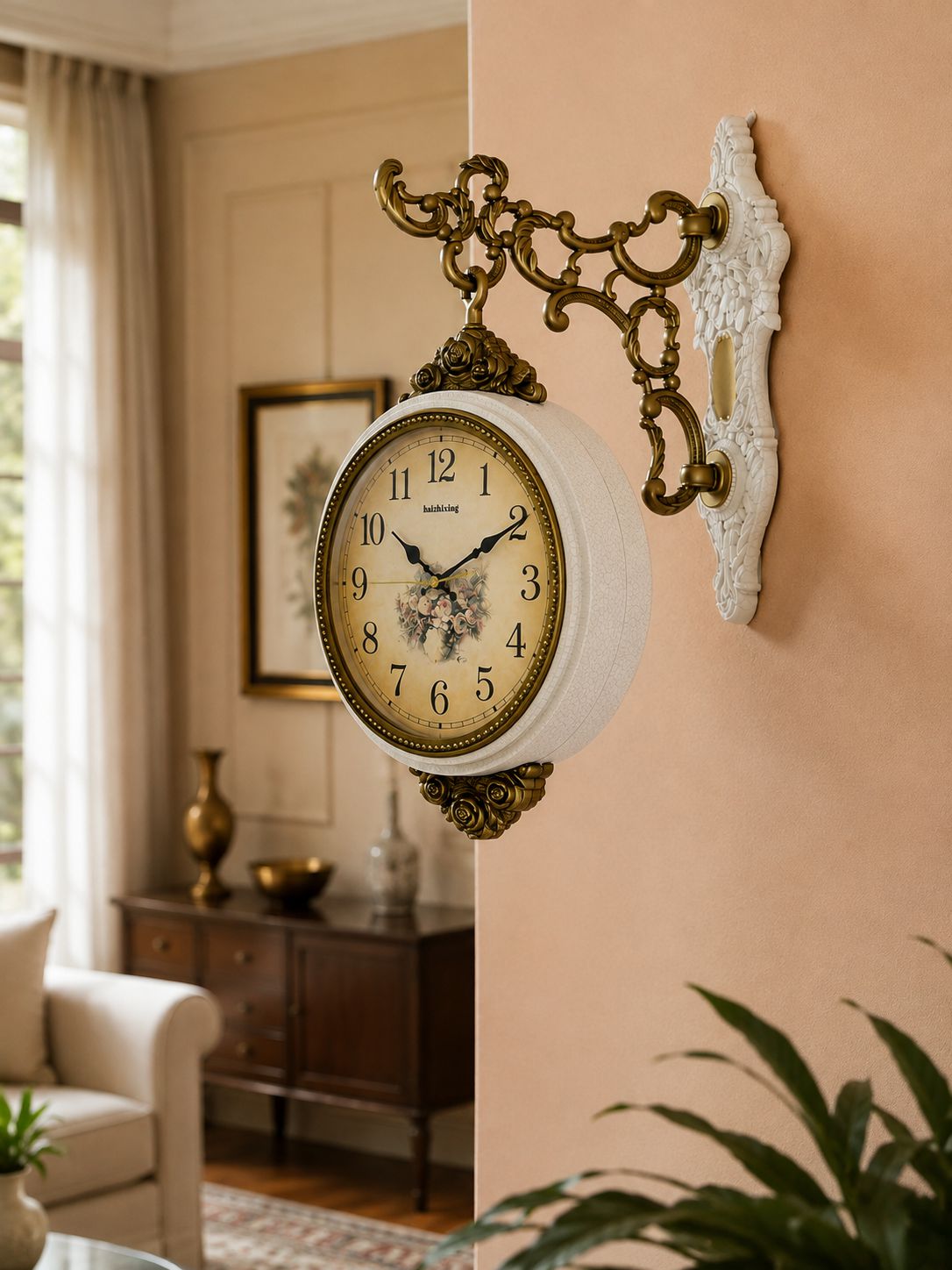 A vintage-style wall clock with an ornate white and gold-toned metal bracket, mounted on a peach-colored wall in a softly lit living room.