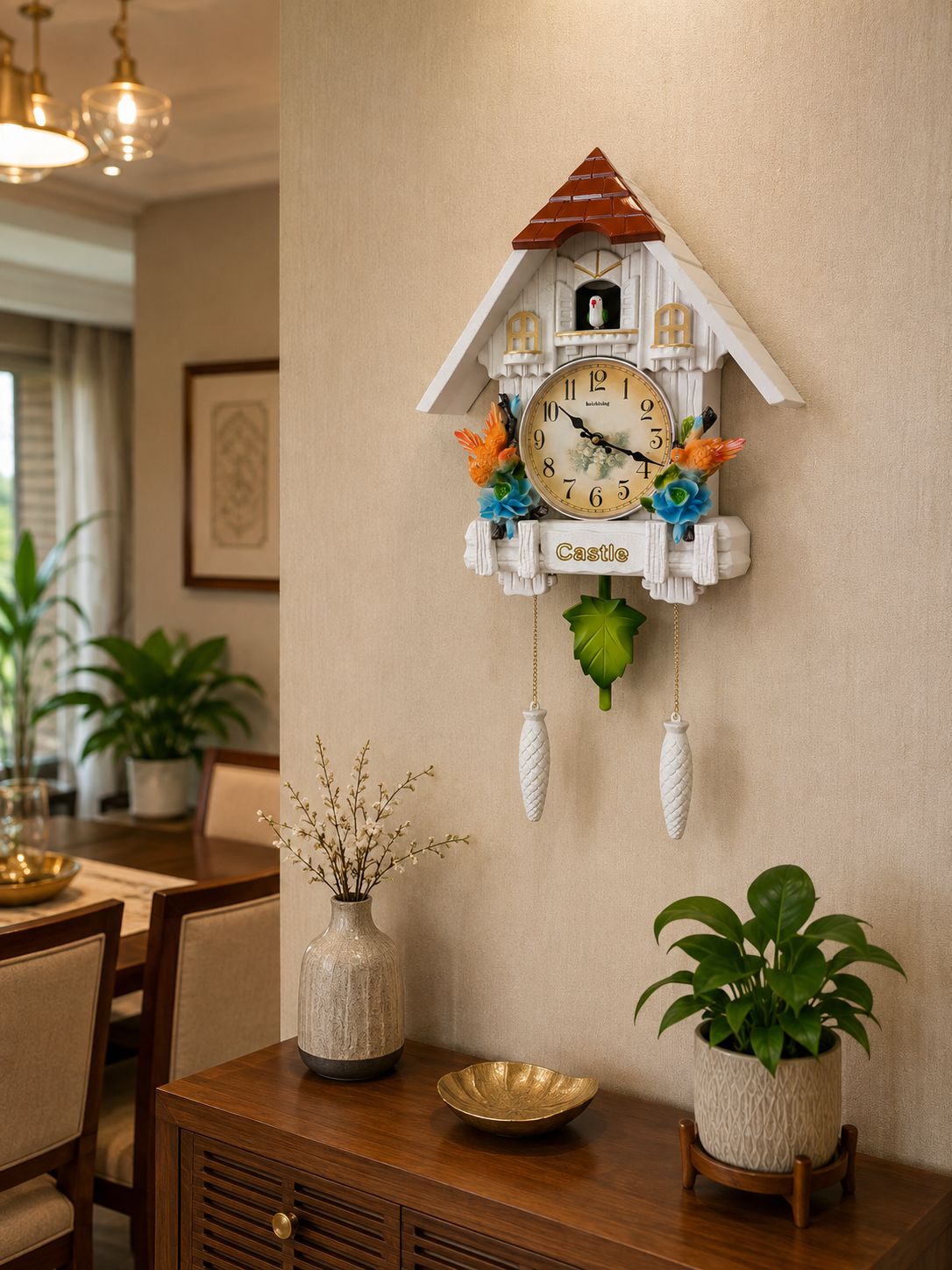 A white cuckoo clock styled like a house with a brown roof, gold-toned clock face, and colorful decorative flowers, mounted on a beige wall in a dining room above a wooden console table with potted plants.