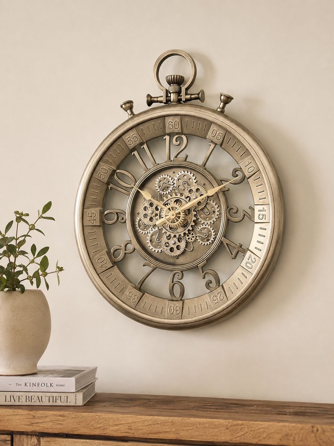 An antique-style wall clock shaped like a pocket watch hangs on a beige wall above a wooden console table. The clock features a distressed bronze finish, skeletonized dial showing intricate inner gears, and raised Arabic numerals.