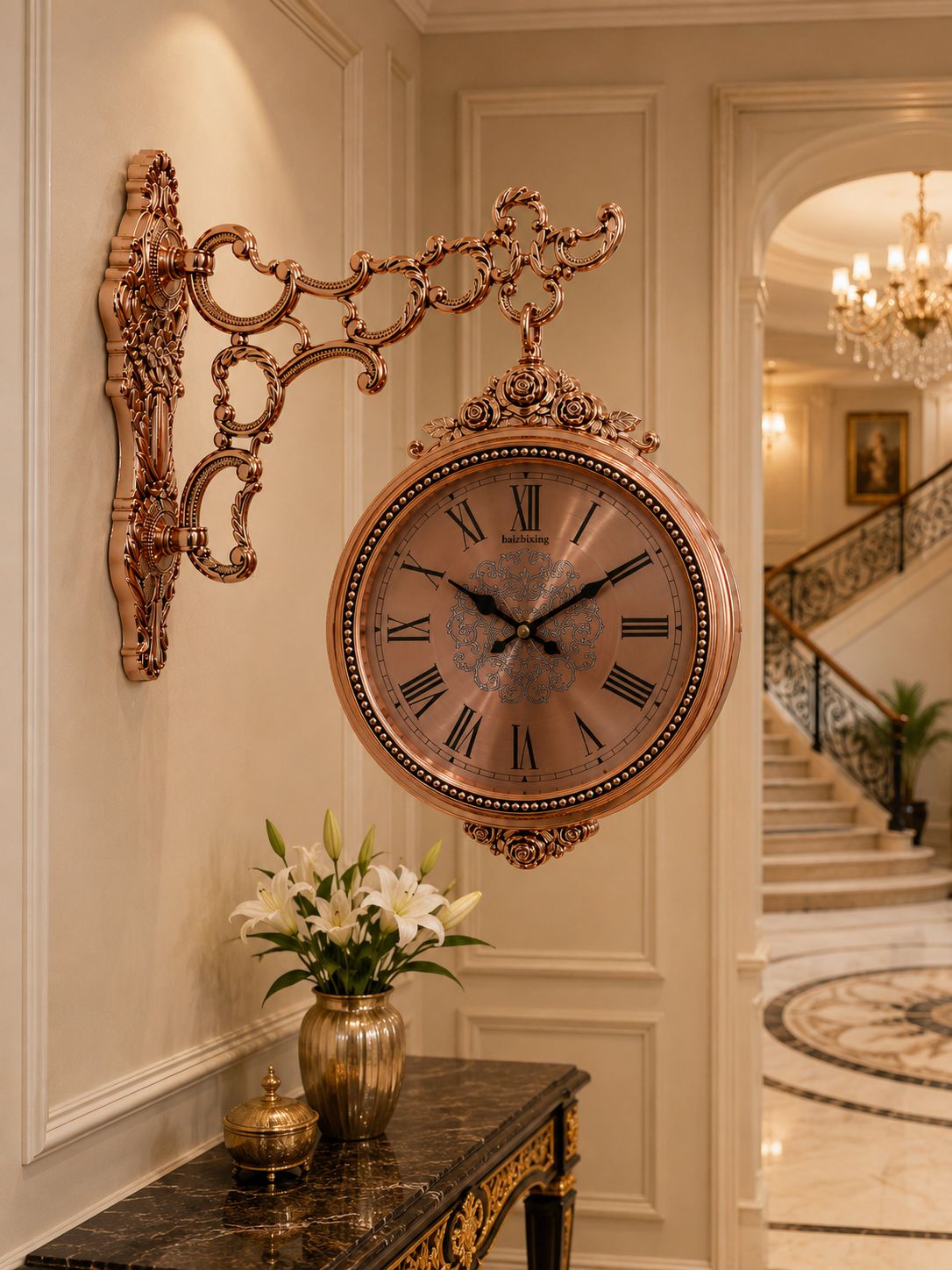 An ornate bronze-finished wall clock with a decorative scroll bracket and a round clock face, mounted on a cream paneled wall in an elegant interior hallway with a console table and flowers below.