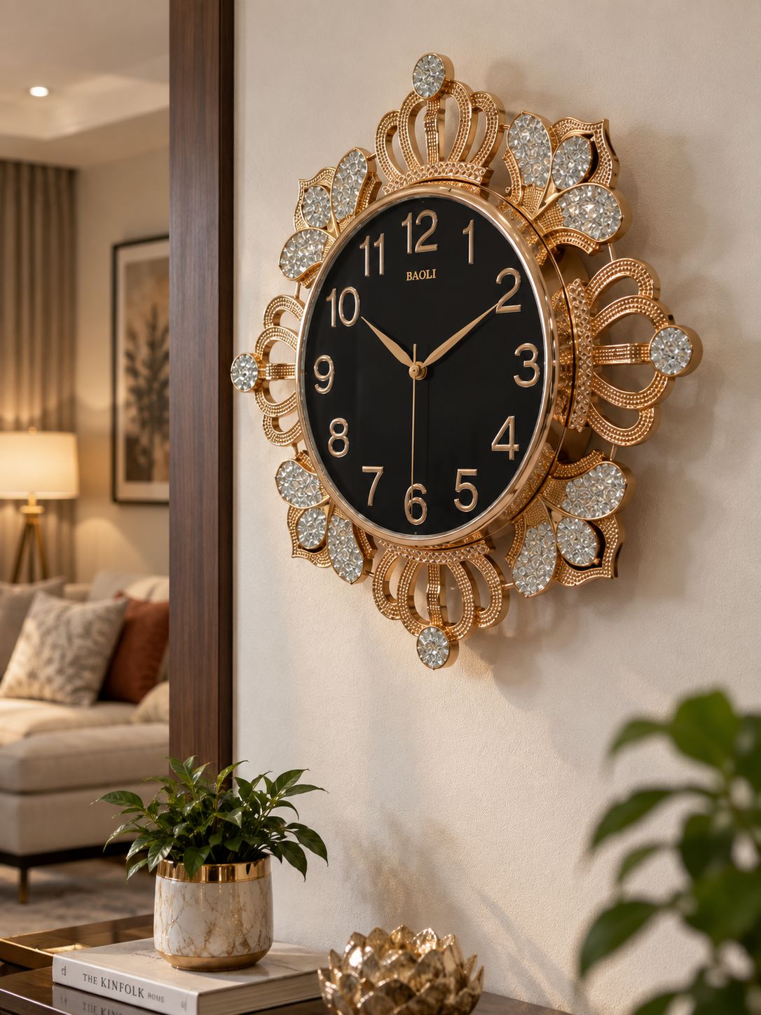 An ornate gold wall clock with a black face and crystal-like embellishments, mounted on a beige wall in a styled living room setting.
