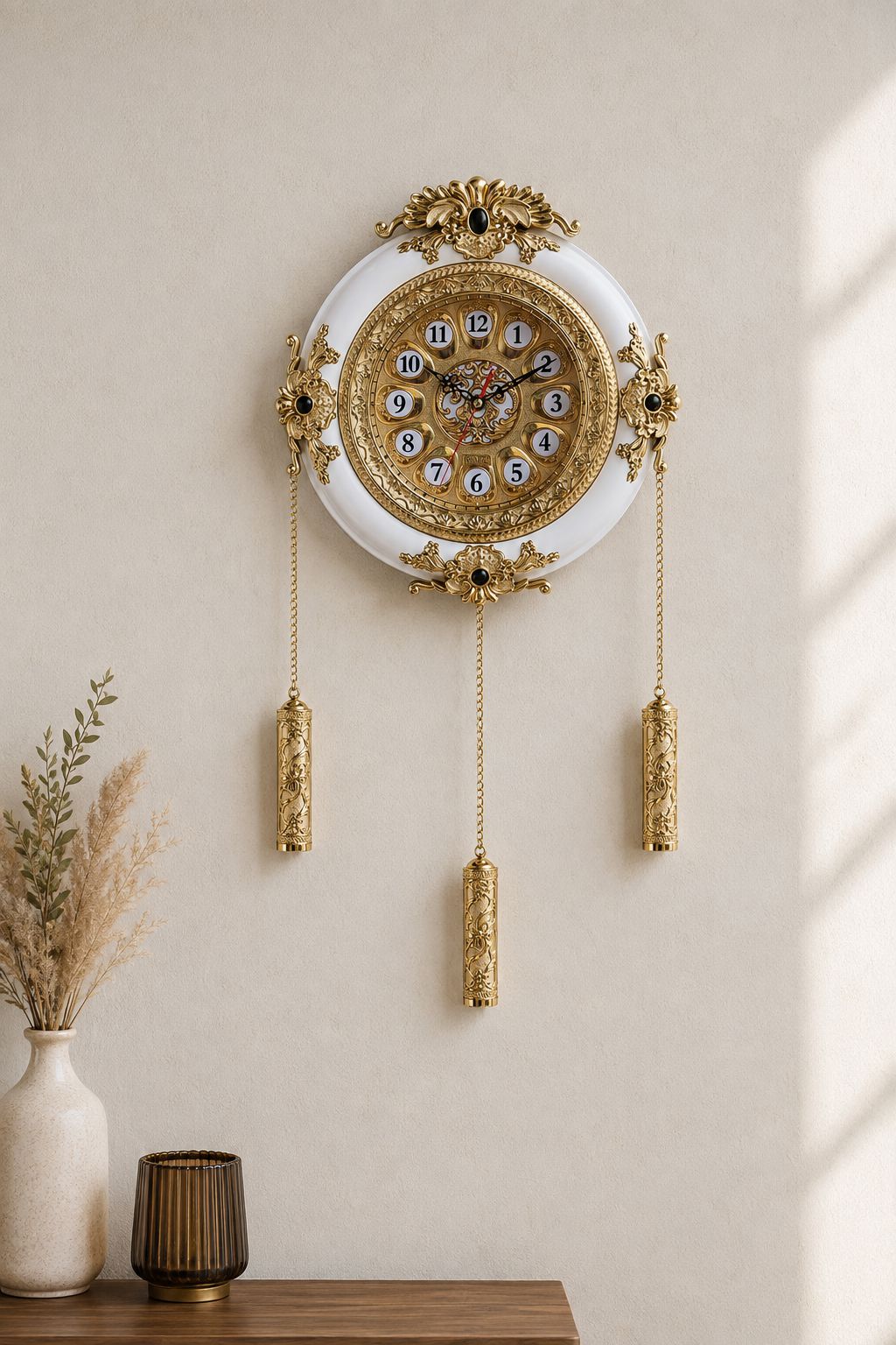 An ornate round wall clock with a white face, gold filigree detailing, and three dangling gold weights, mounted on a beige wall above a wooden surface with decorative vases.