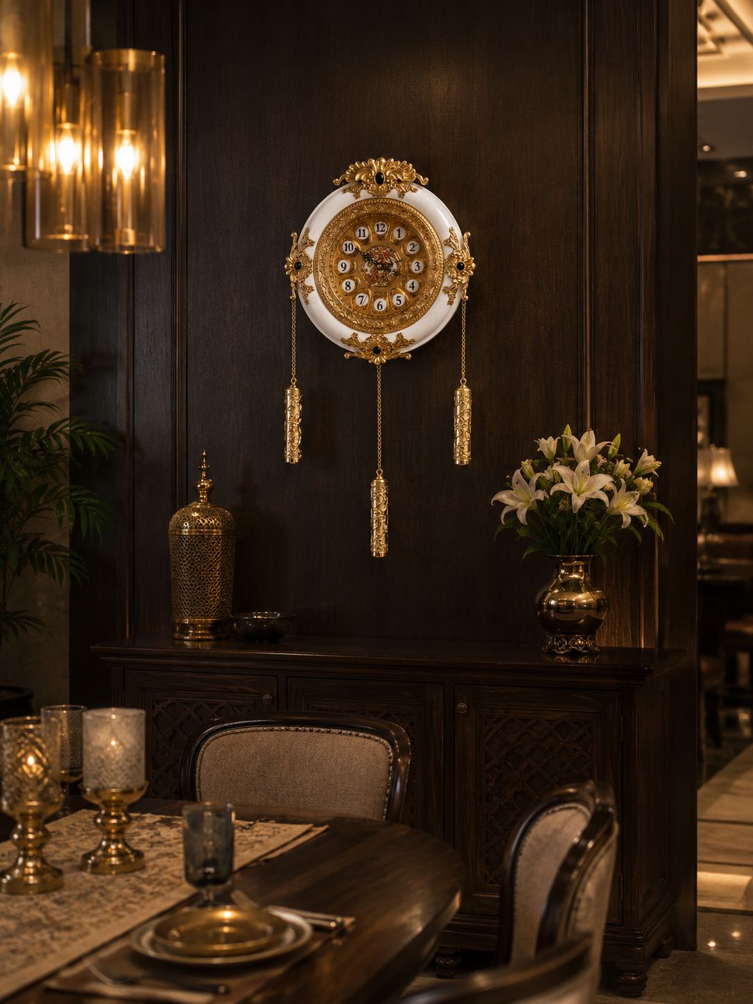 An ornate round wall clock with gold and white detailing hangs on a dark wood-paneled wall above a matching dark wooden console table. The scene is styled as a luxury dining room with a dining table, chairs, a vase of white flowers, and warm hanging lights.