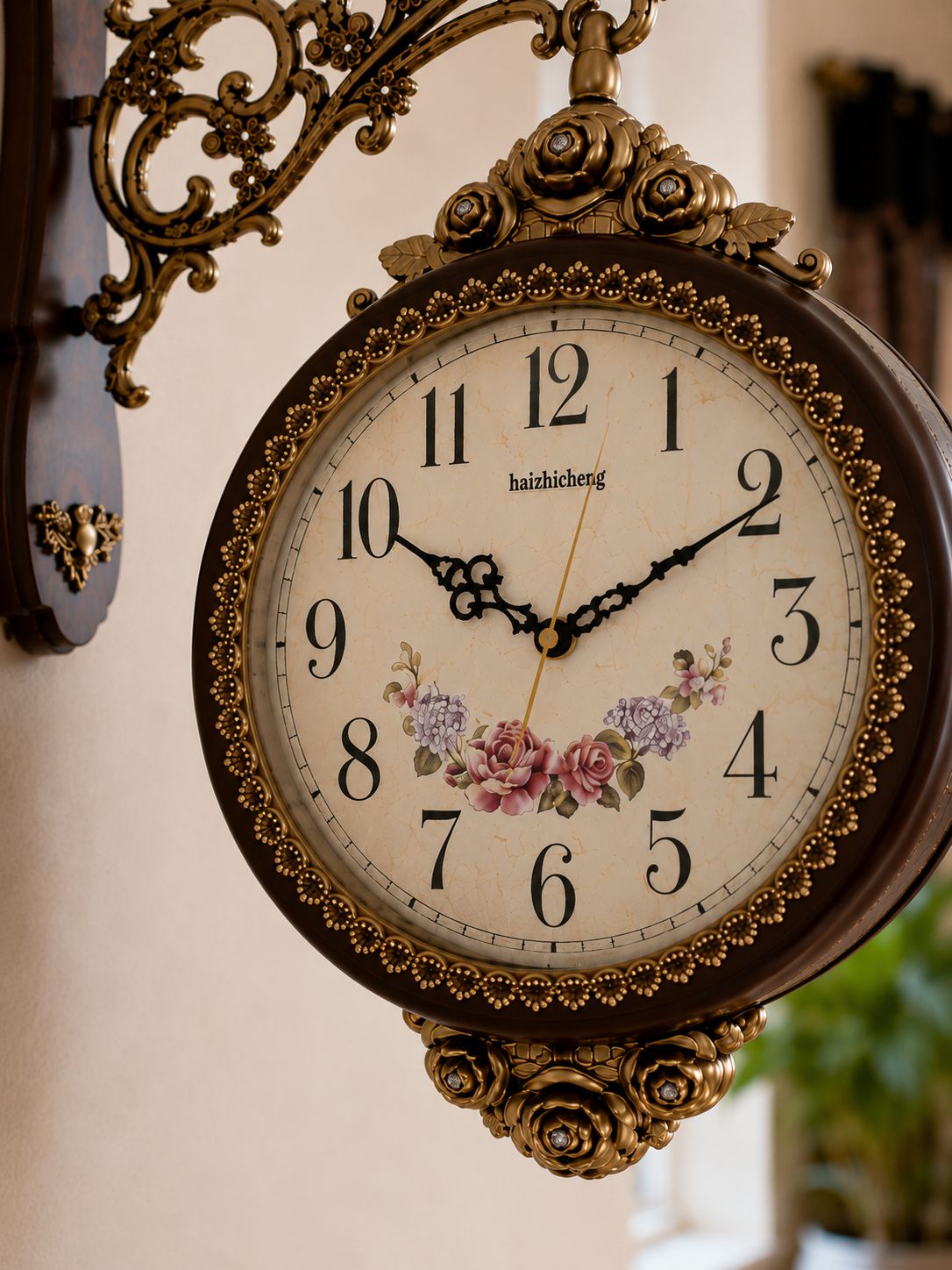 An ornate vintage-style wall clock with a dark brown frame, gold-tone metal scrollwork mount, and a cream dial decorated with black Arabic numerals, floral artwork, and the brand name 'haizhicheng'.