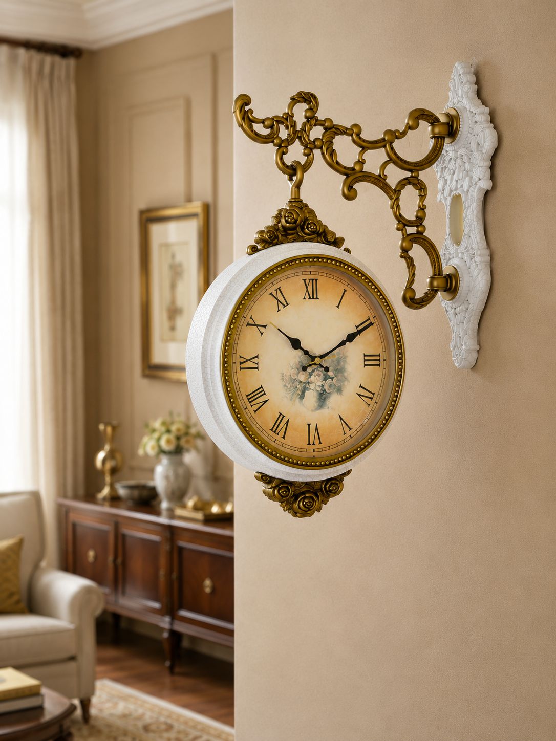 An ornate vintage-style wall clock with a white body, gold-tone scrollwork bracket, and Roman numeral face, mounted on a beige wall in a furnished room.