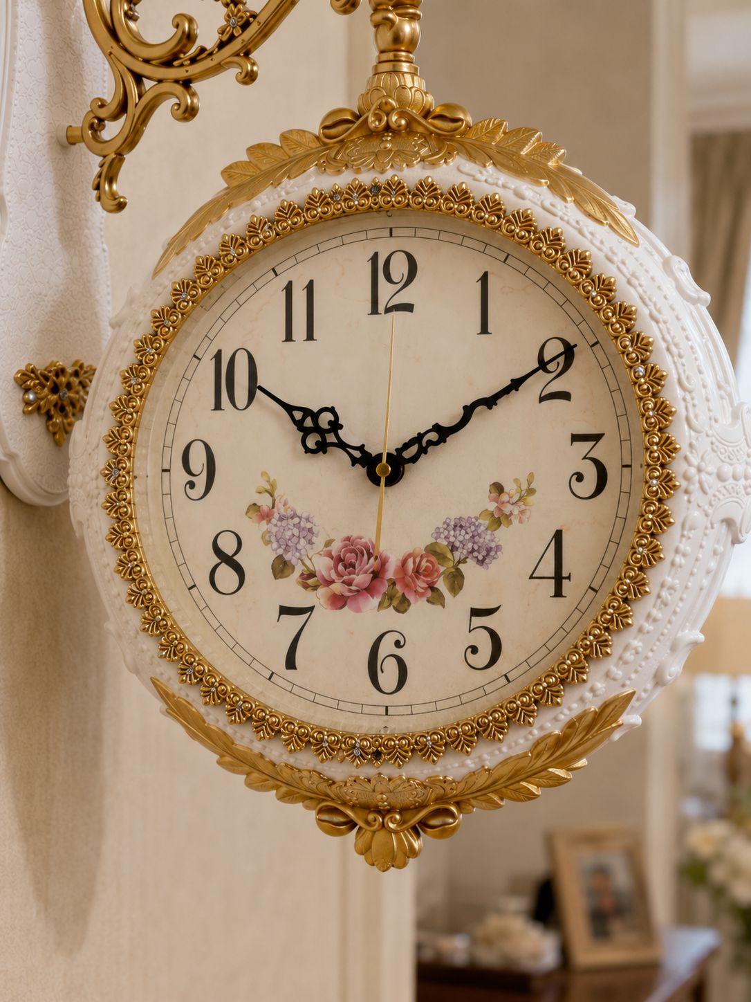 An ornate vintage-style wall clock with a white lace-embroidered frame, gold gilded detailing, black Arabic numerals, and delicate pink and purple floral illustrations on the clock face, shown hanging in an elegant interior setting.