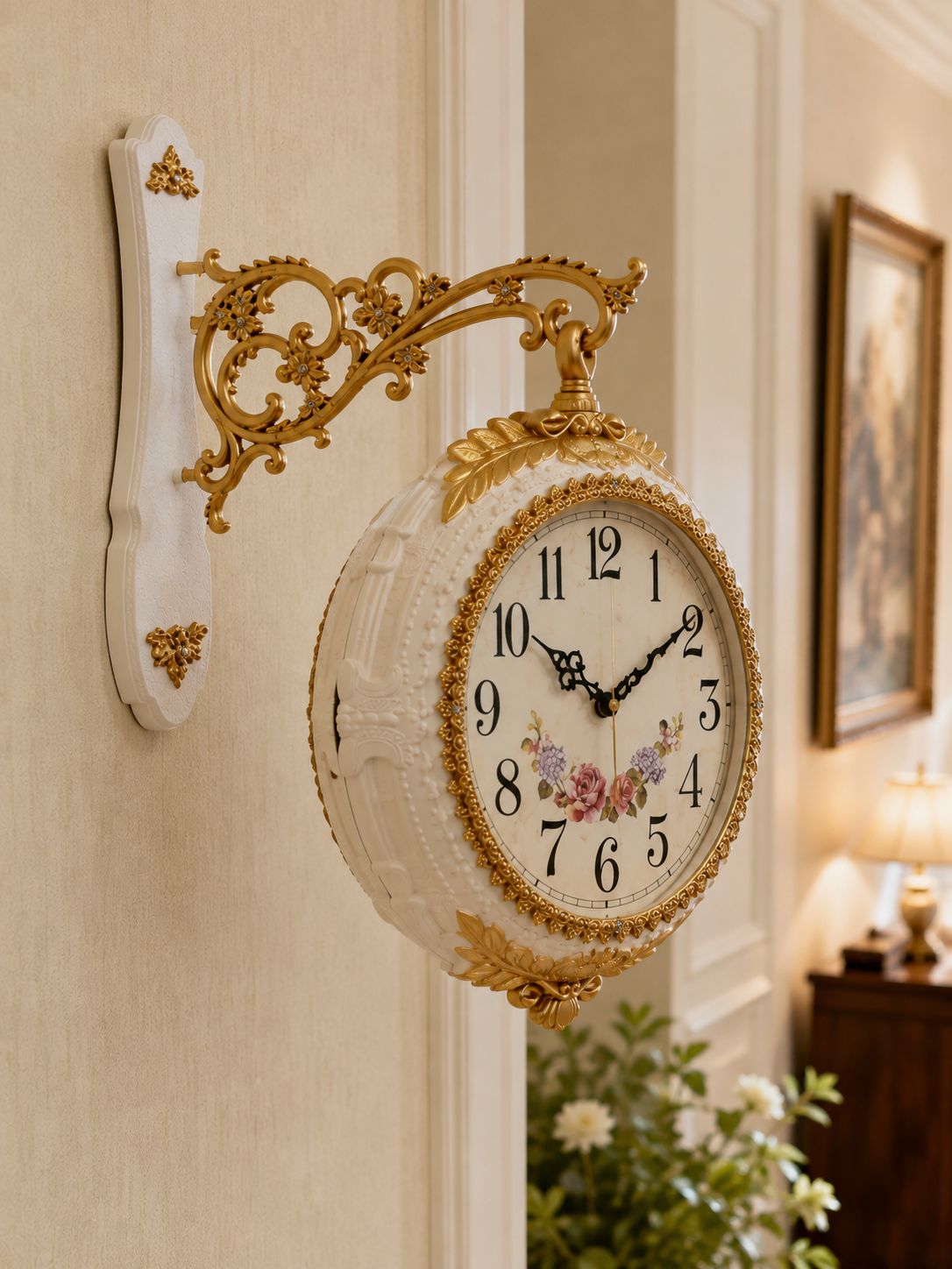 An ornate vintage-style wall clock with a white round body, gold scrollwork bracket, floral clock face, and black numerals, mounted on a beige wall in an elegant interior setting.