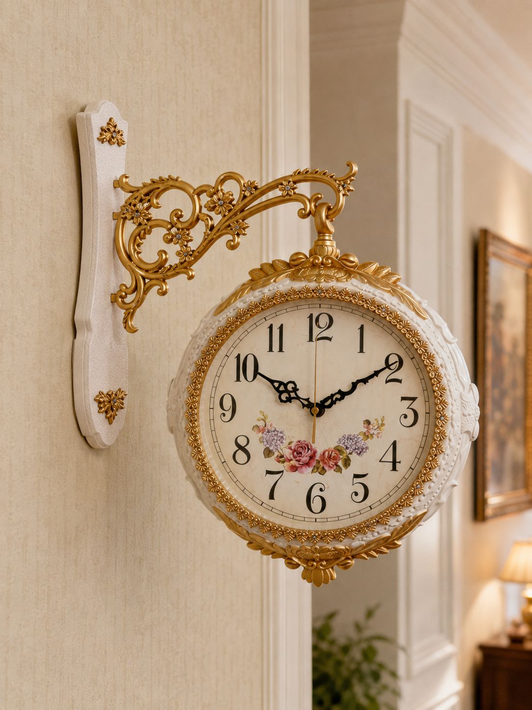 An ornate wall-mounted clock with a white carved frame, gold scrollwork bracket, and floral details on the clock face, shown hanging on a beige wall in an indoor hallway setting.