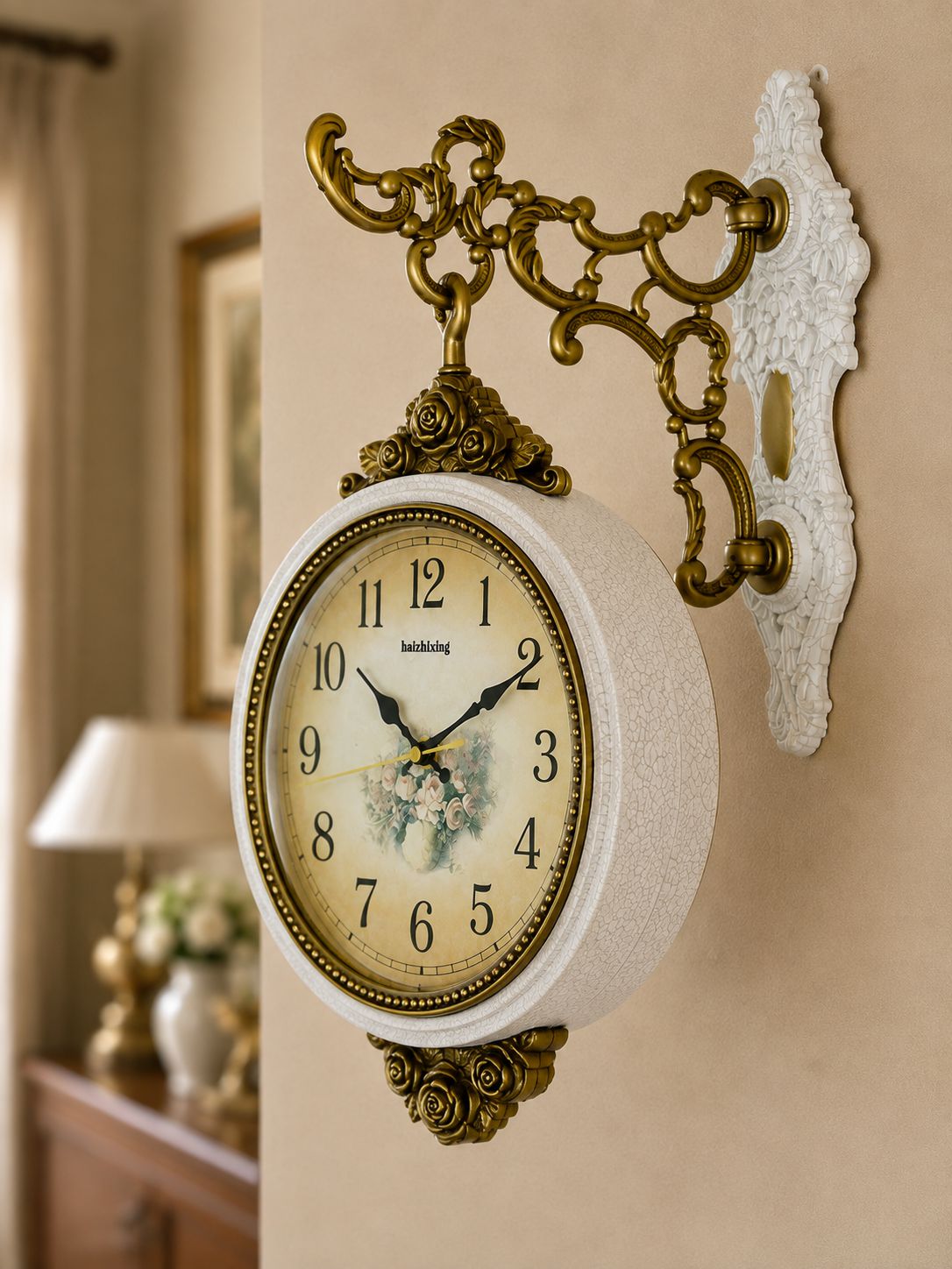 An ornate wall clock with a round cream dial, black Roman numerals, and a floral illustration at the center, mounted on a beige wall with a decorative gold bracket and white wall plate.