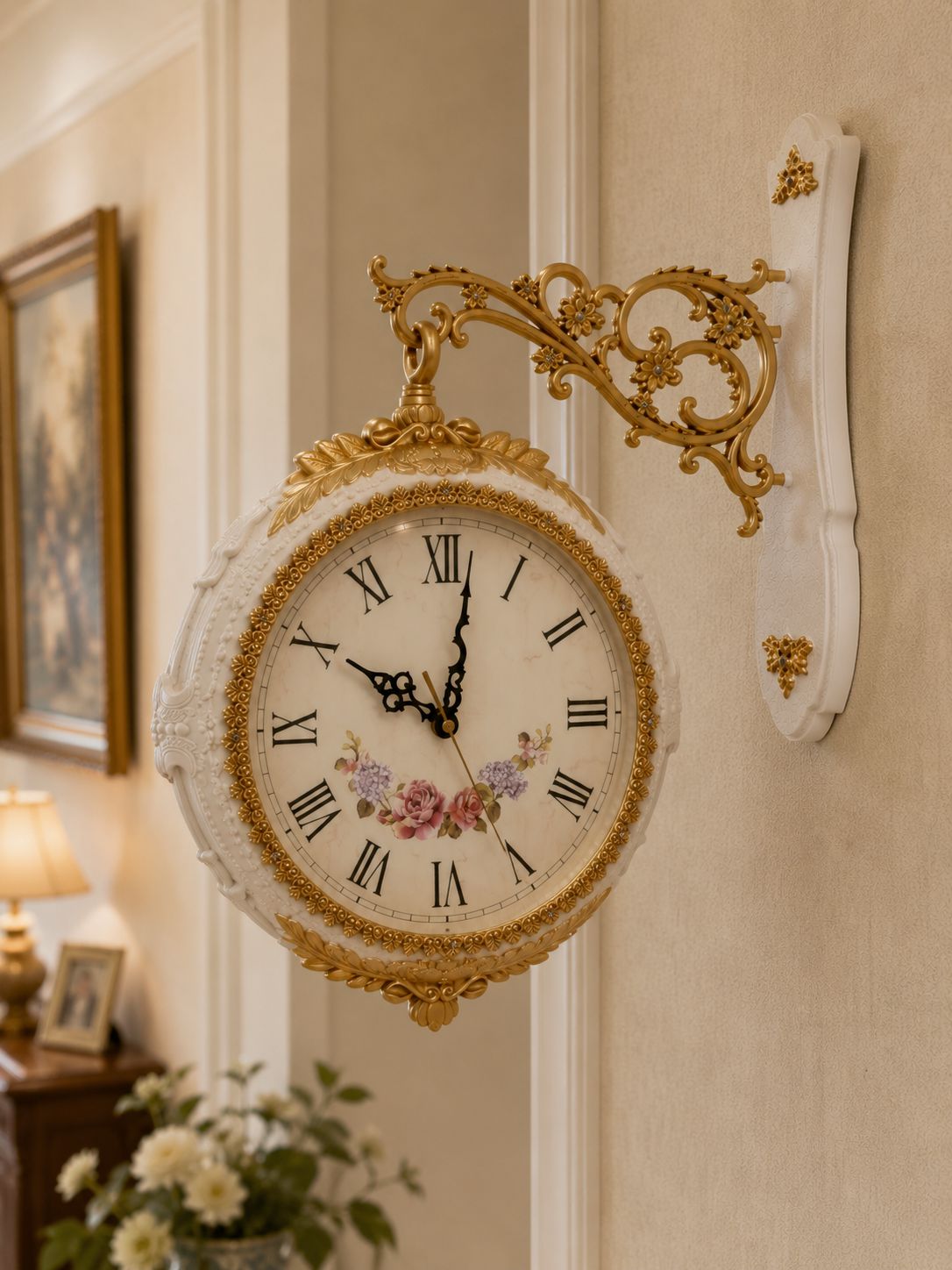 An ornate wall clock with a white carved frame, gold scrollwork bracket, black Roman numerals, and a floral illustration on the clock face, mounted on a beige wall in an elegant interior setting.