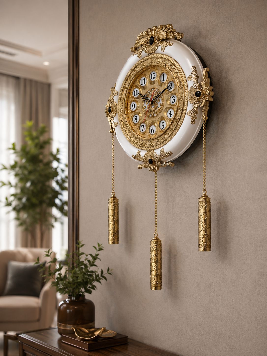 An ornate wall clock with a white oval face, gold decorative detailing, and three hanging gold cylinder weights, mounted on a beige wall in a styled interior room.