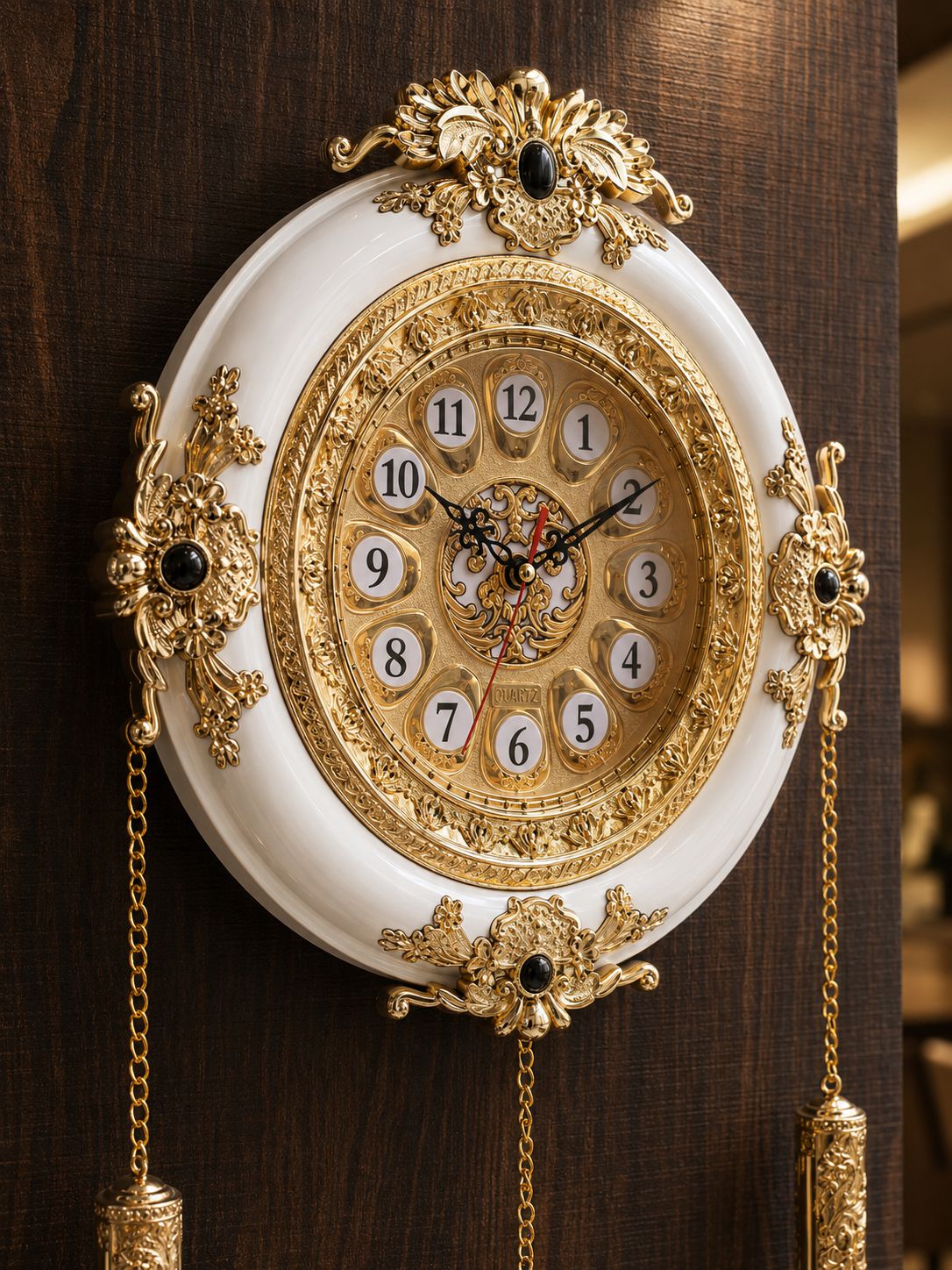 An ornate wall clock with a white oval face, intricate gold filigree detailing, gold chains, and black decorative accents, mounted on a dark wood panel.