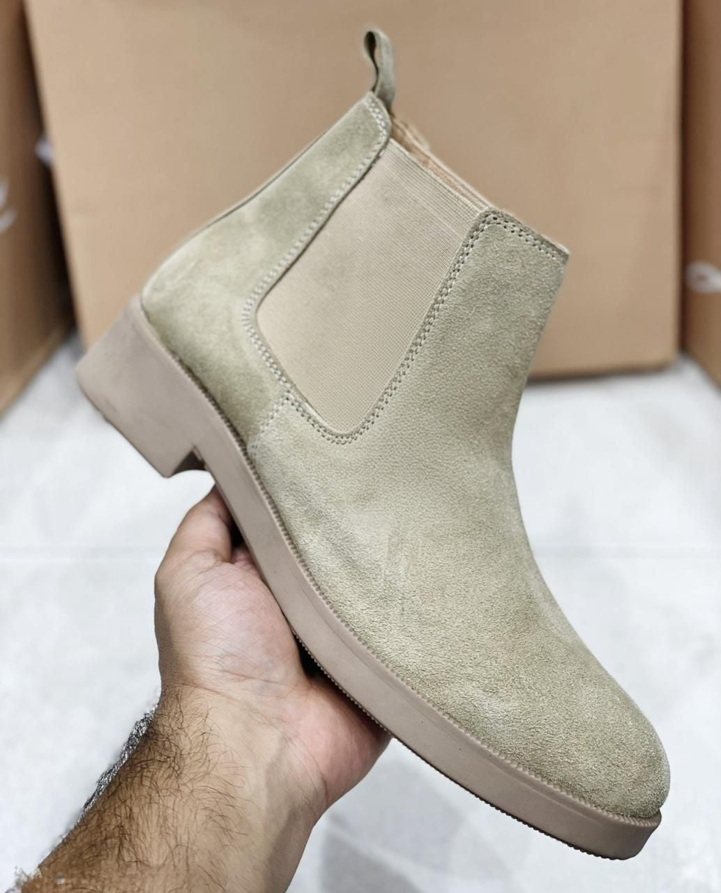 Beige suede ankle boot held by a hand with a cardboard box in the background